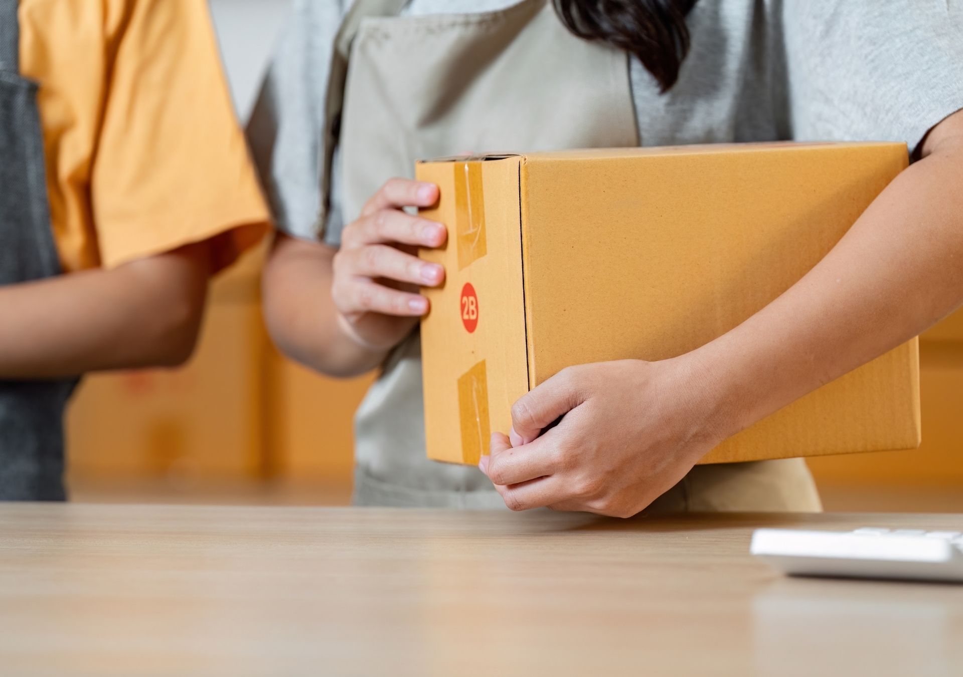 Two people holding a brown cardboard box on a wooden table, likely for shipping.