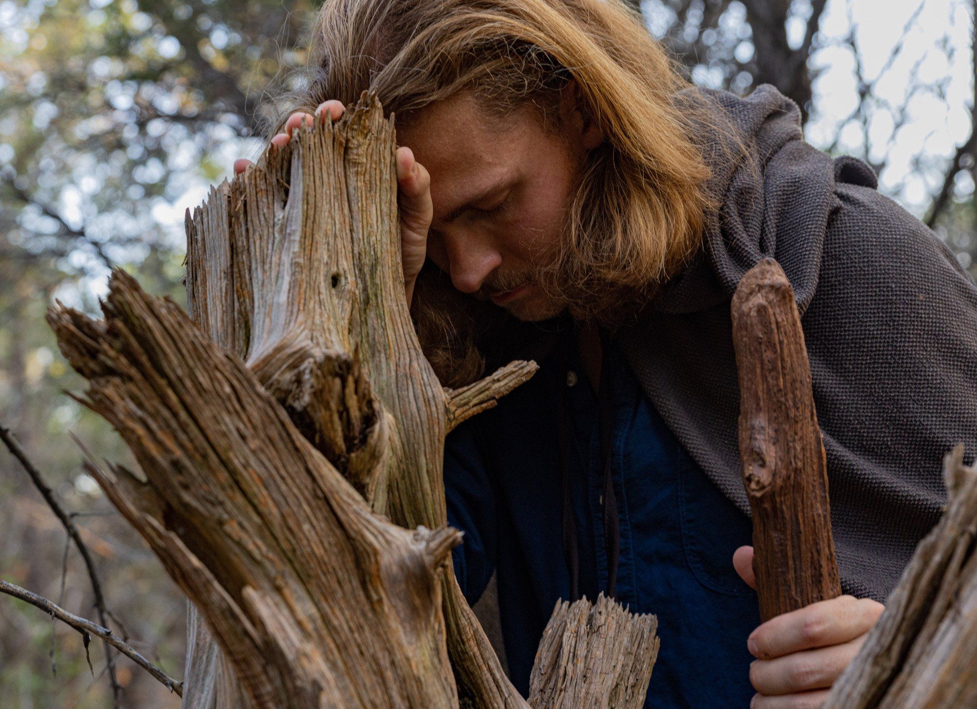 Man with long hair, leaning head on weathered wood. Wearing a cloak, in a forest.