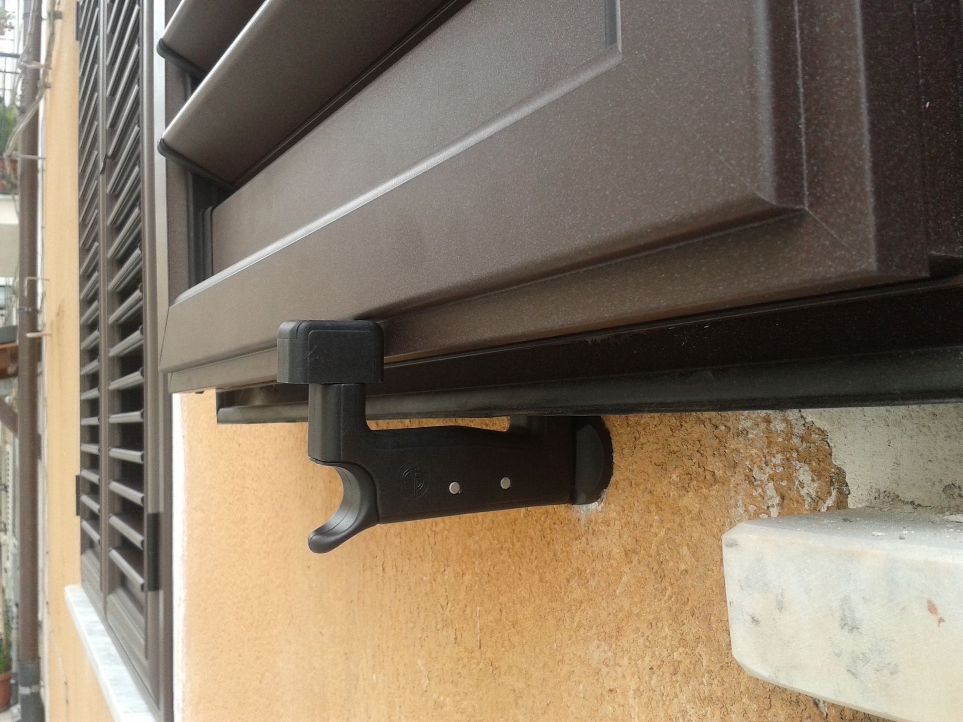 Un gancio di bloccaggio nero per persiana, montato su una parete gialla con superficie ruvida, sotto una persiana marrone.