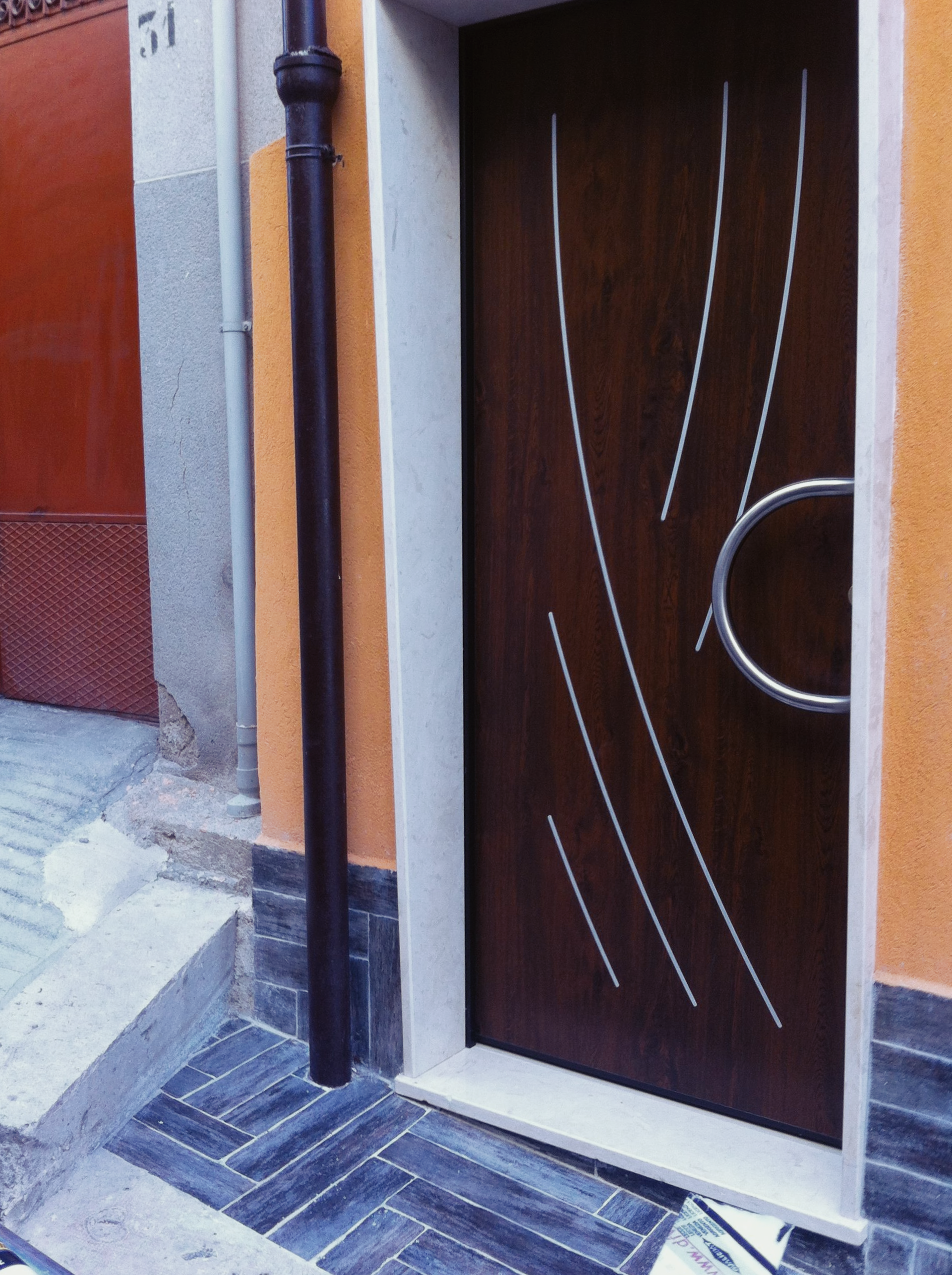 Una porta d'ingresso in legno scuro con dettagli argentati curvi e una maniglia circolare, incastonata in una parete arancione chiaro con battiscopa piastrellato.