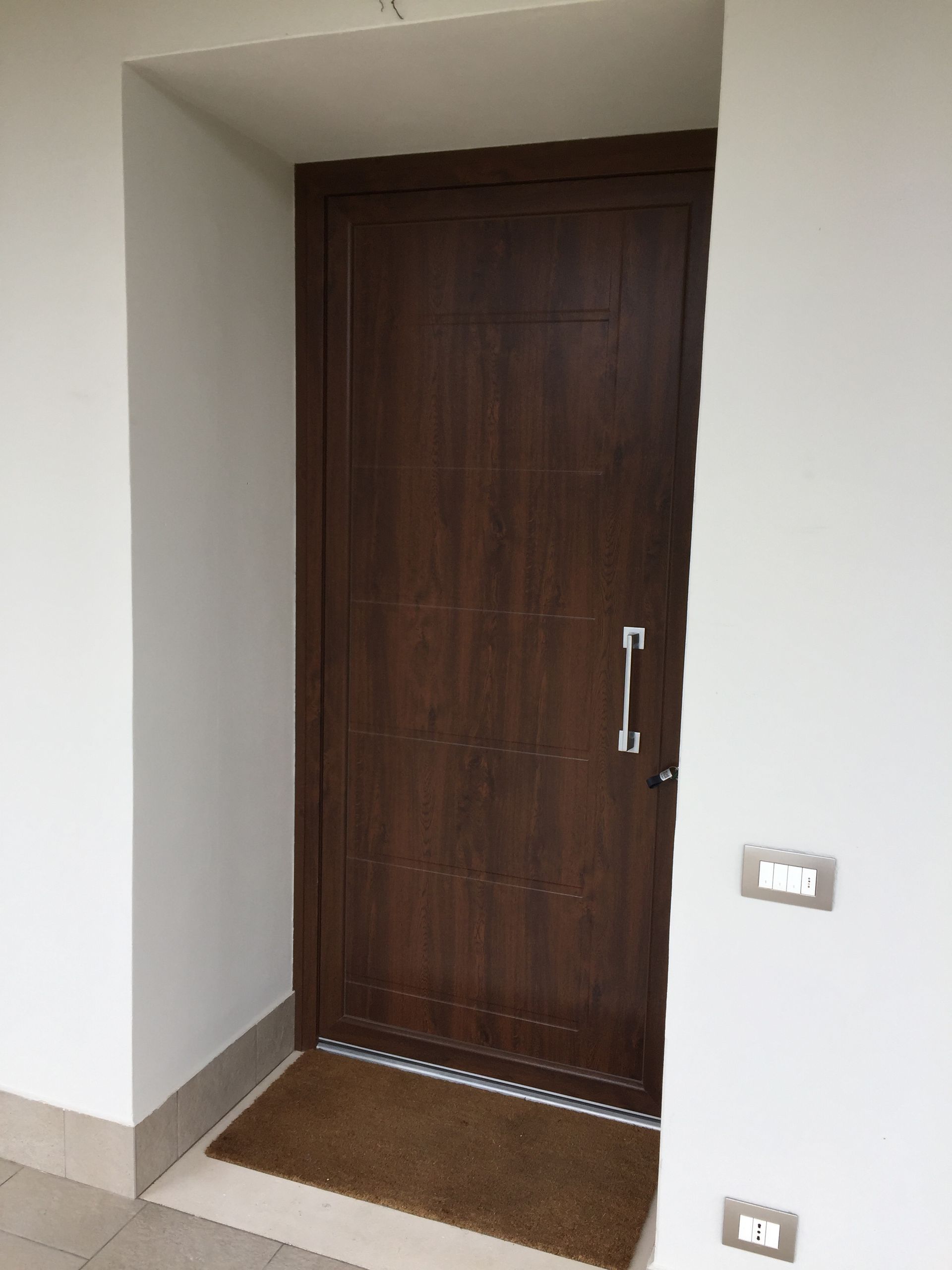 Una porta di legno marrone scuro con una maniglia verticale argentata, incastonata in una parete bianca sopra un piccolo zerbino marrone.