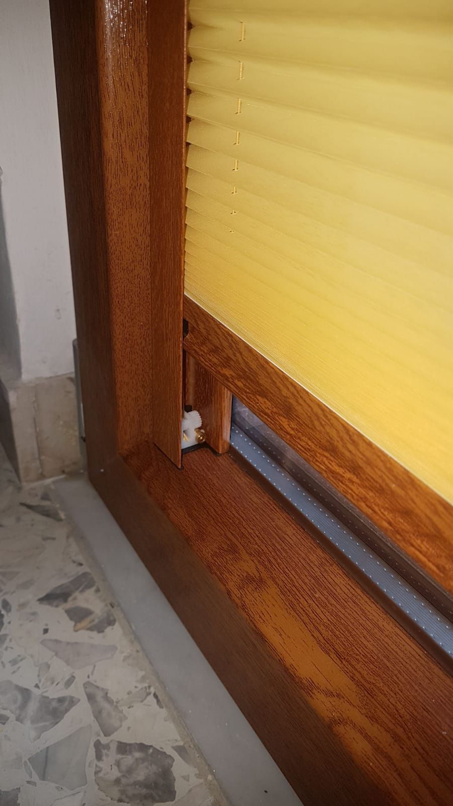 Primo piano di un telaio di finestra in legno con una tenda a pieghe gialla, che mostra una piccola staffa di montaggio in plastica bianca.