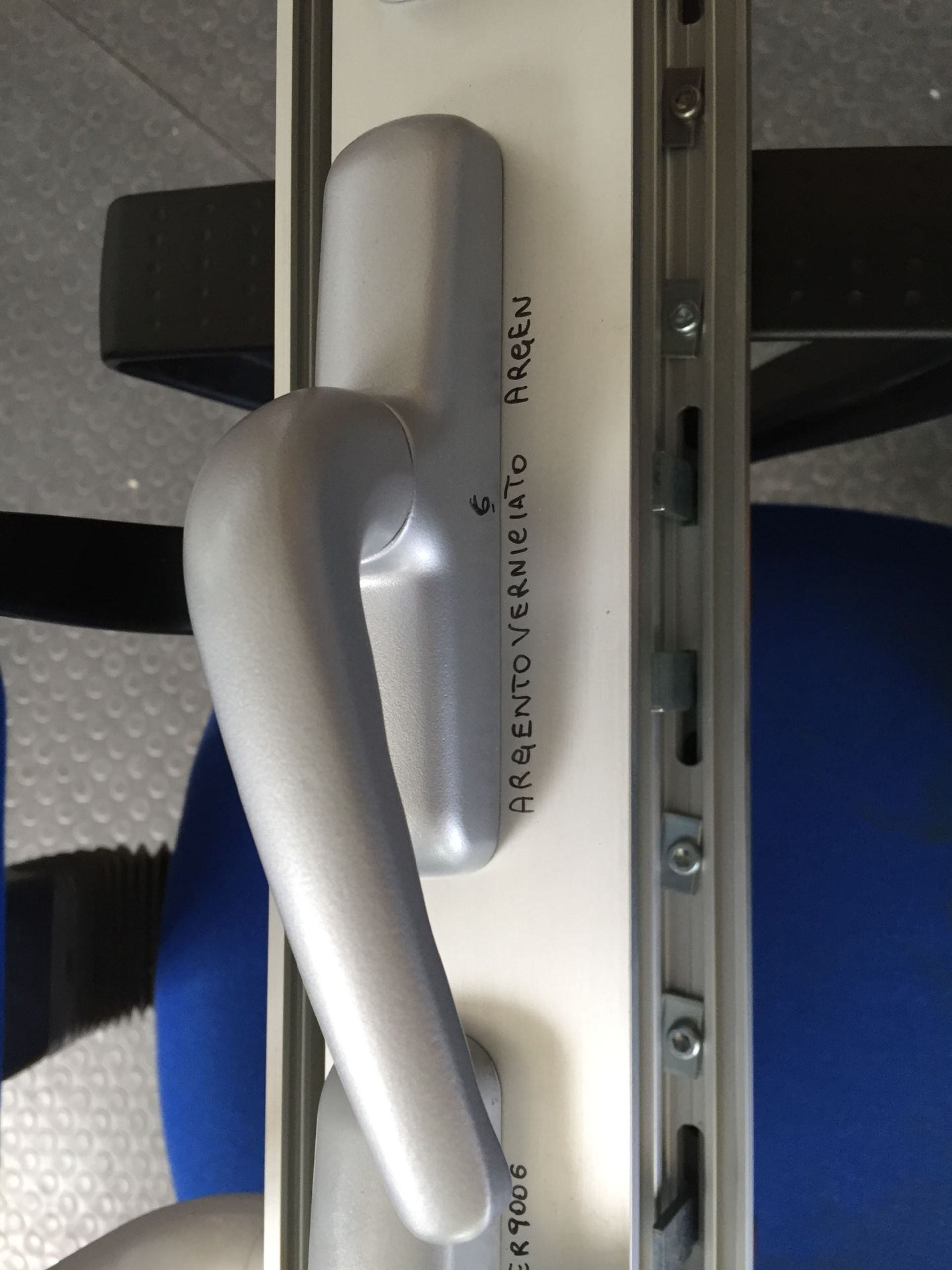 Una maniglia per finestra color argento montata su una cornice bianca, che mostra parte del meccanismo di chiusura con punti di aggancio in metallo.