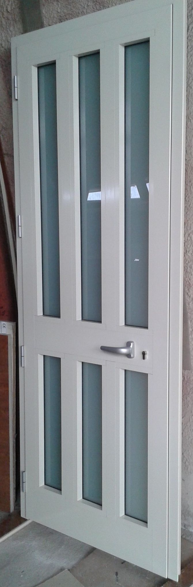 Una porta interna singola bianca con sei pannelli verticali in vetro satinato e maniglia argentata, mostrata addossata a una parete.