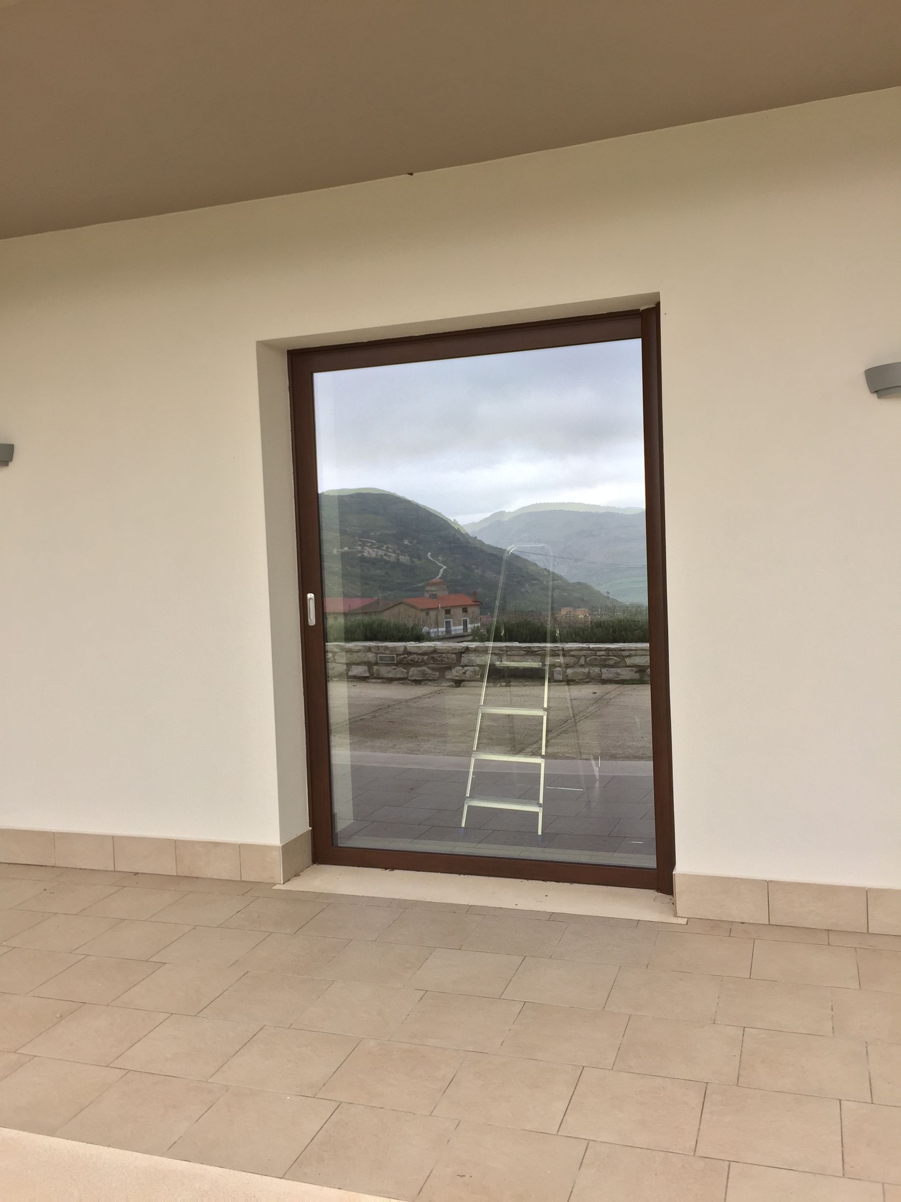 Una porta scorrevole in vetro con cornice marrone, vista da un patio, riflette una scala e un paesaggio montano in lontananza.