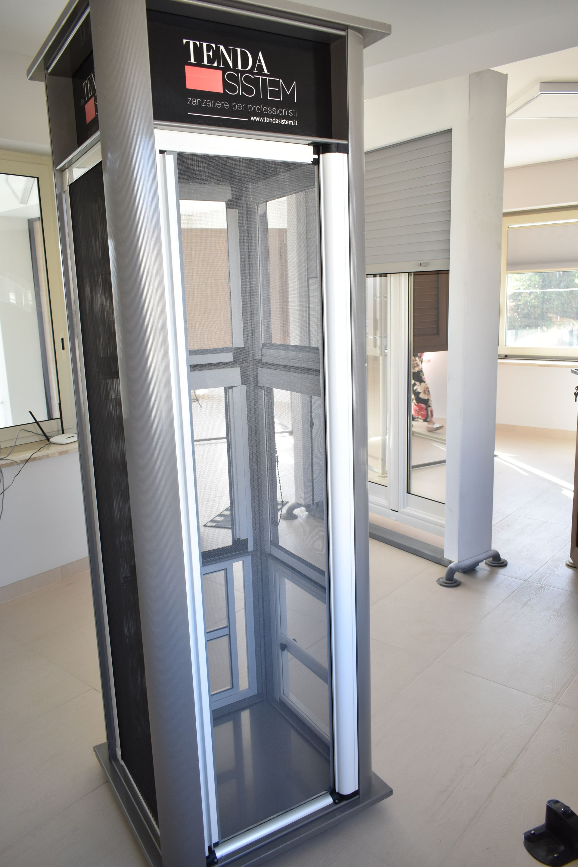 Un modello da esposizione argentato, da terra, di una zanzariera o tenda Tenda System, in un luminoso e moderno showroom.