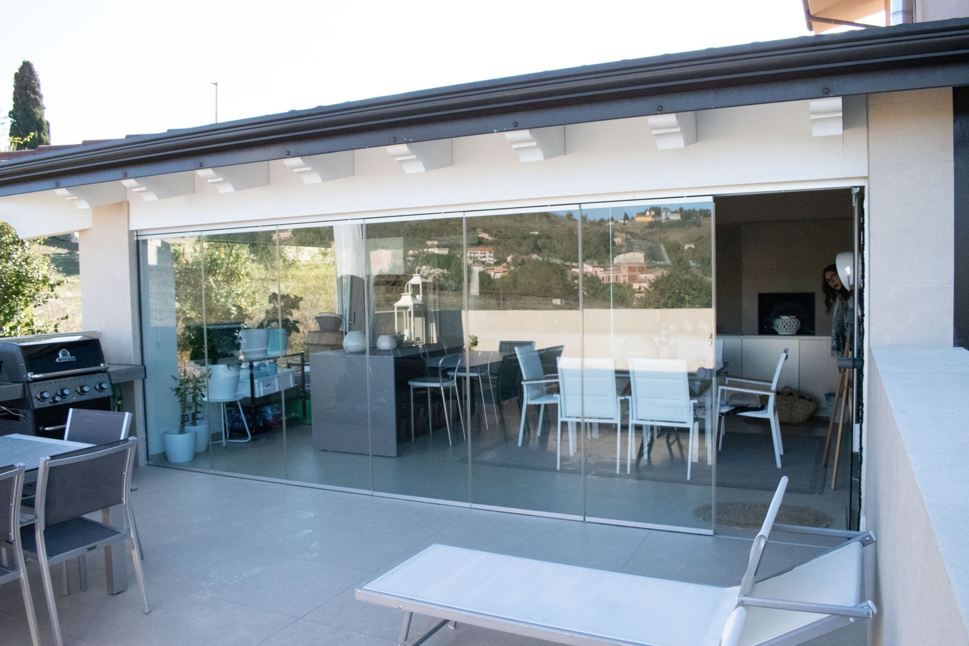 Un patio esterno vetrato con mobili da pranzo e barbecue, visibile da una terrazza piastrellata di colore chiaro.