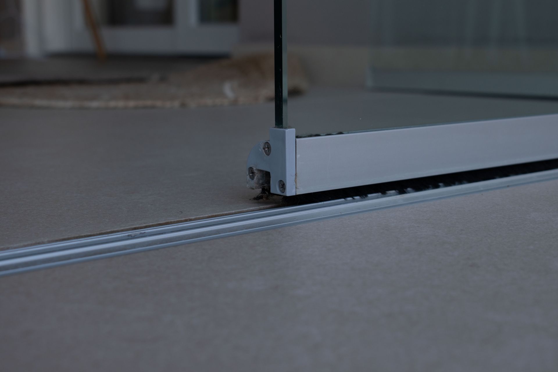 Primo piano della base e del meccanismo di scorrimento di una porta scorrevole in vetro argentata, incastonata in un pavimento piastrellato grigio chiaro.