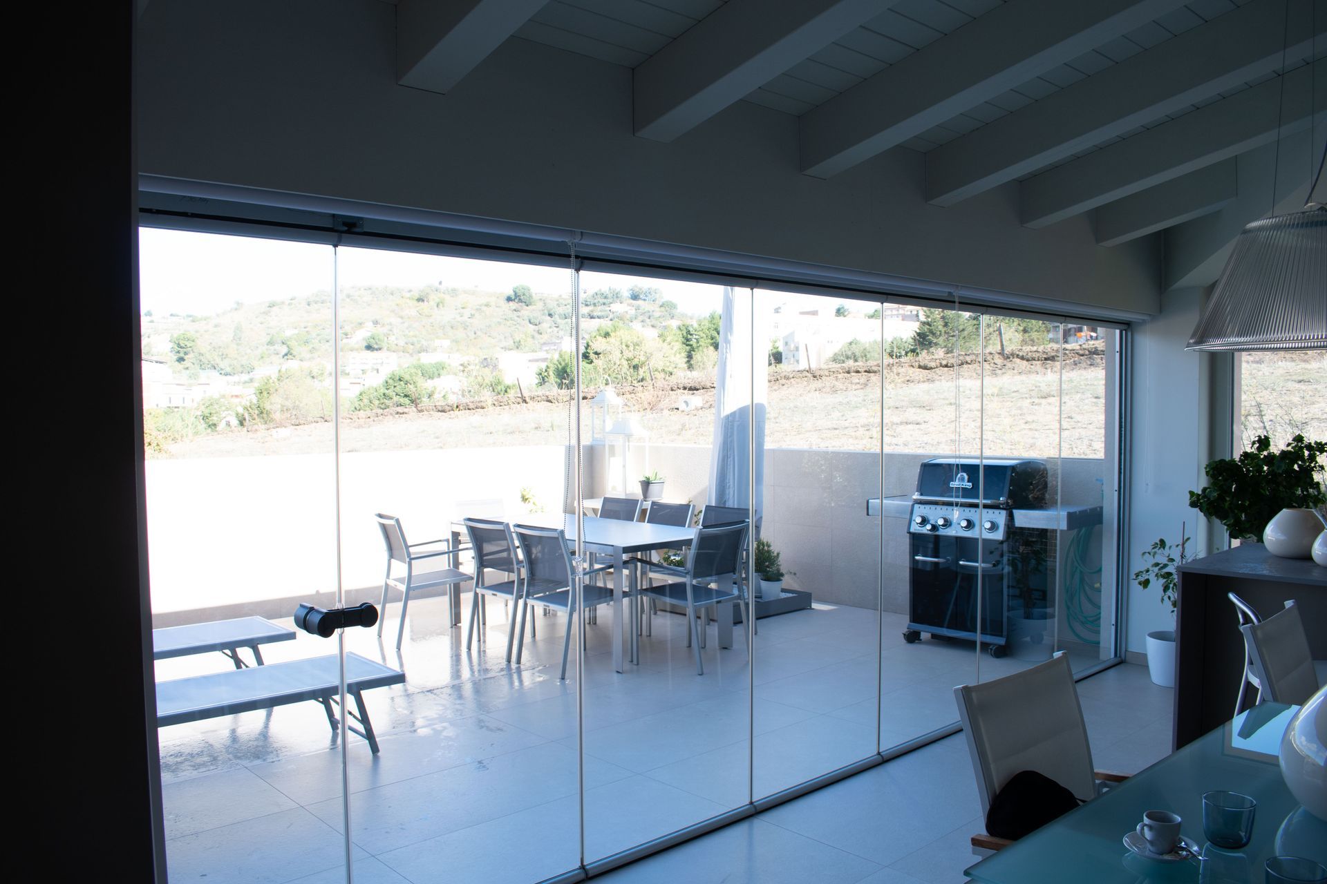 Un moderno patio coperto con pareti vetrate a tutta altezza, mobili da esterno e barbecue, affacciato su un paesaggio soleggiato.