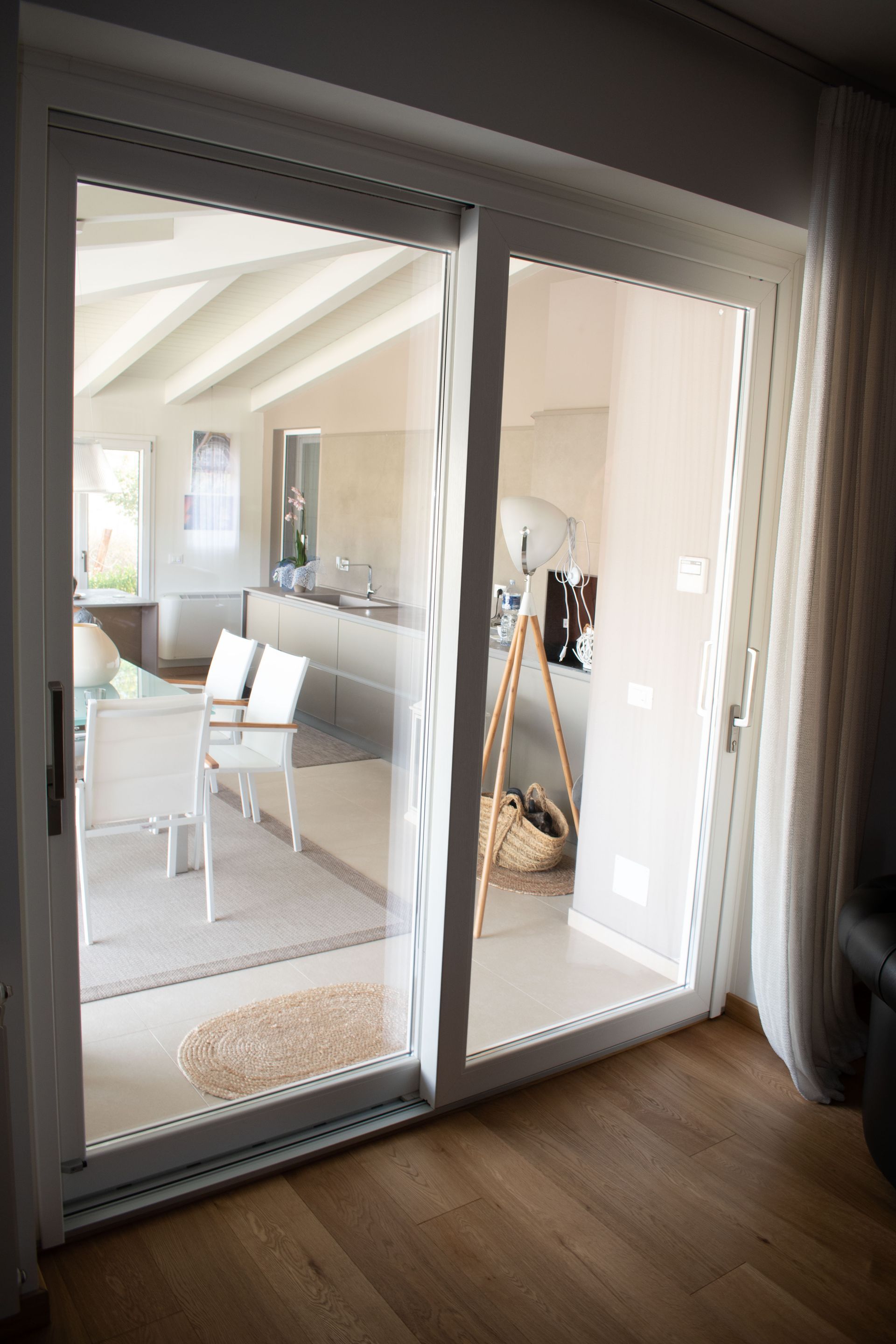 Attraverso un'ampia porta scorrevole in vetro si intravede una sala da pranzo luminosa, arredata con sedie bianche, un tavolo e una lampada da terra.