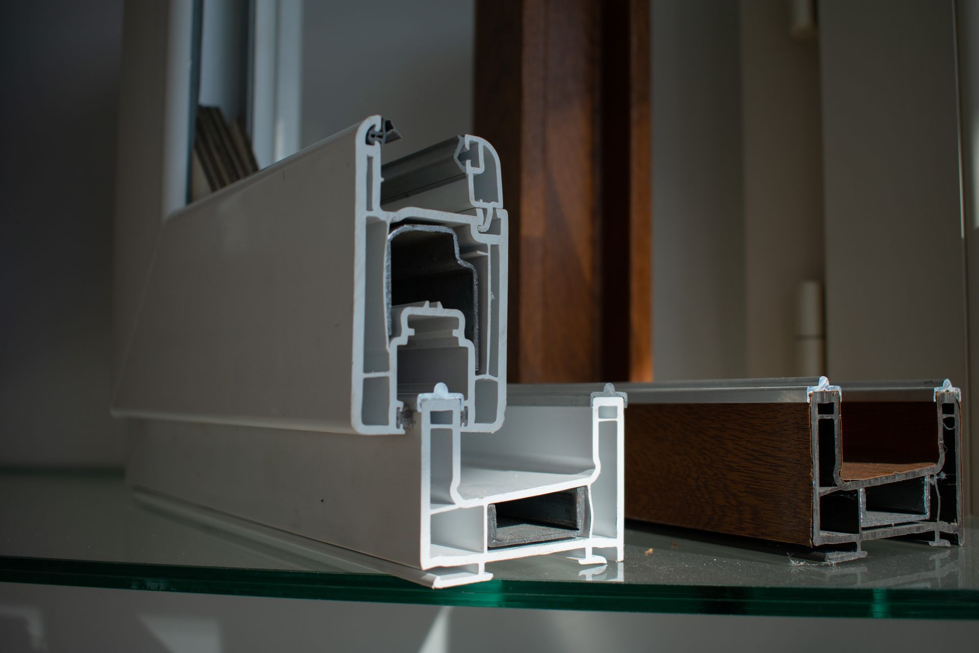 Campioni in sezione trasversale di profili per telai di finestre in PVC bianco e alluminio marrone, esposti su una superficie di vetro.