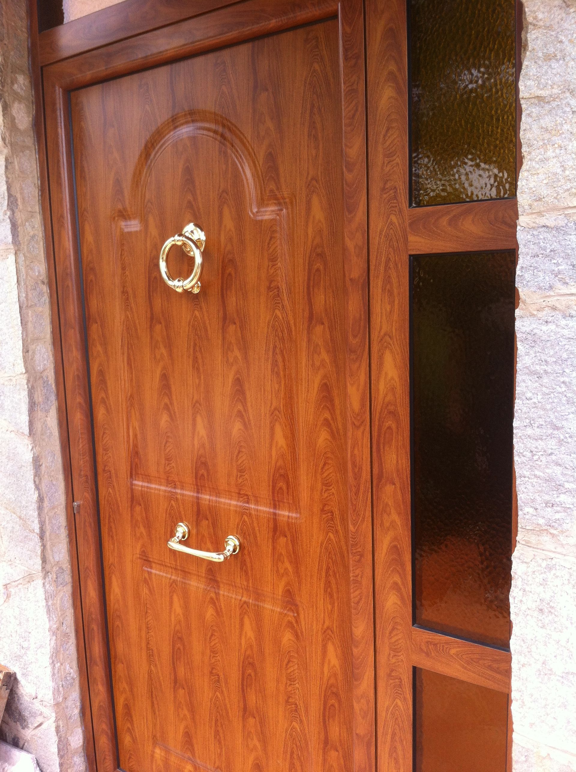 Una porta d'ingresso con finitura effetto legno marrone, battente e maniglia dorati, accanto a una finestra verticale con vetro color ambra.