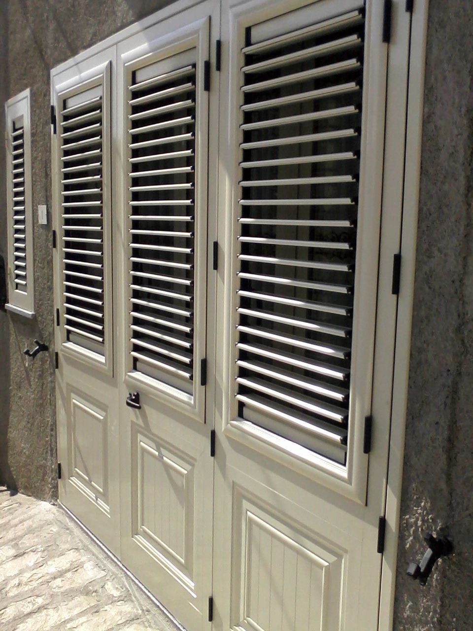 Due doppie porte a pannelli multipli color crema con persiane a lamelle orizzontali, incastonate in un muro esterno in pietra.