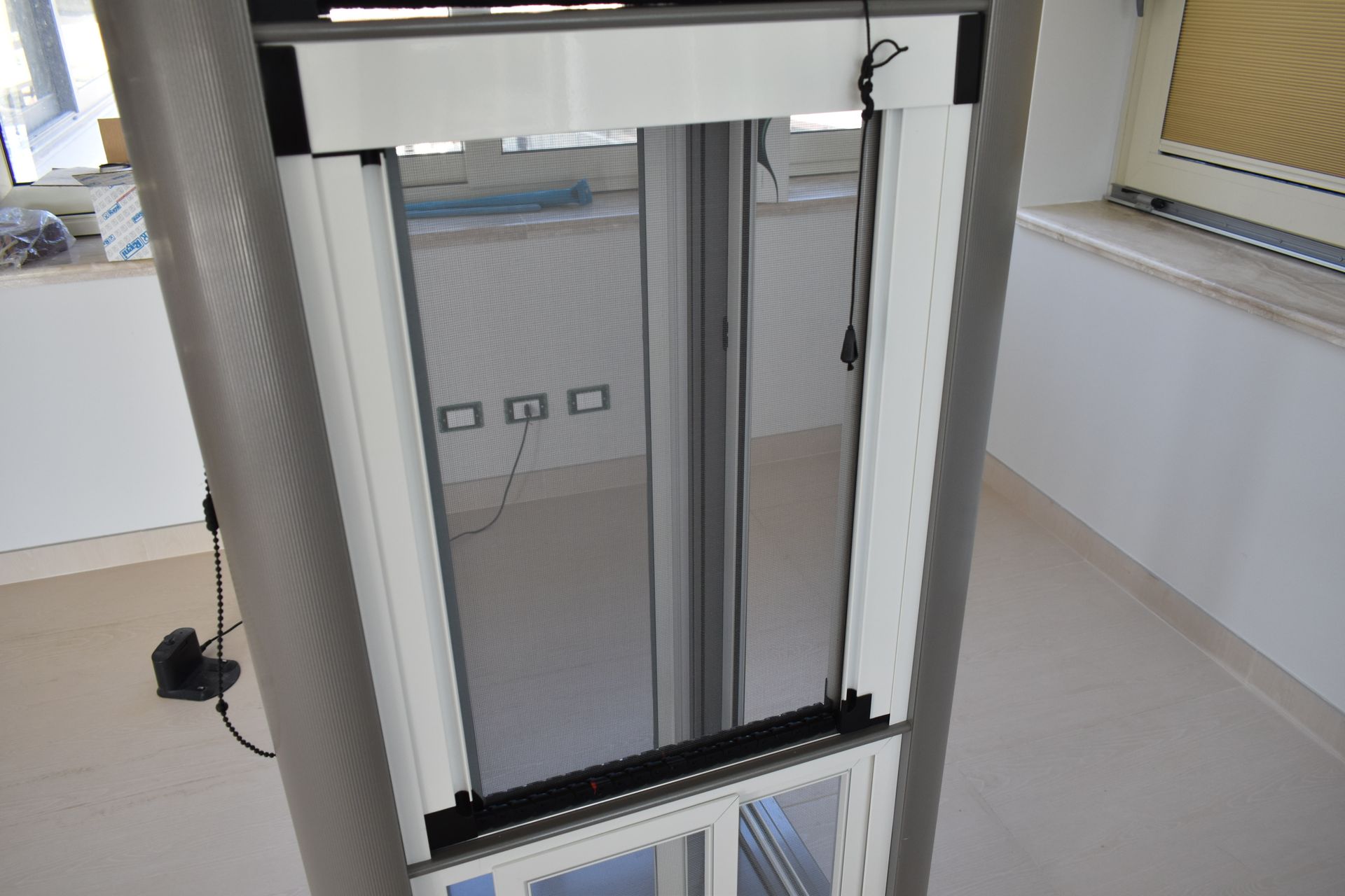 Zanzariera verticale con cornice bianca installata su una finestra in una stanza con pareti chiare e prese elettriche.