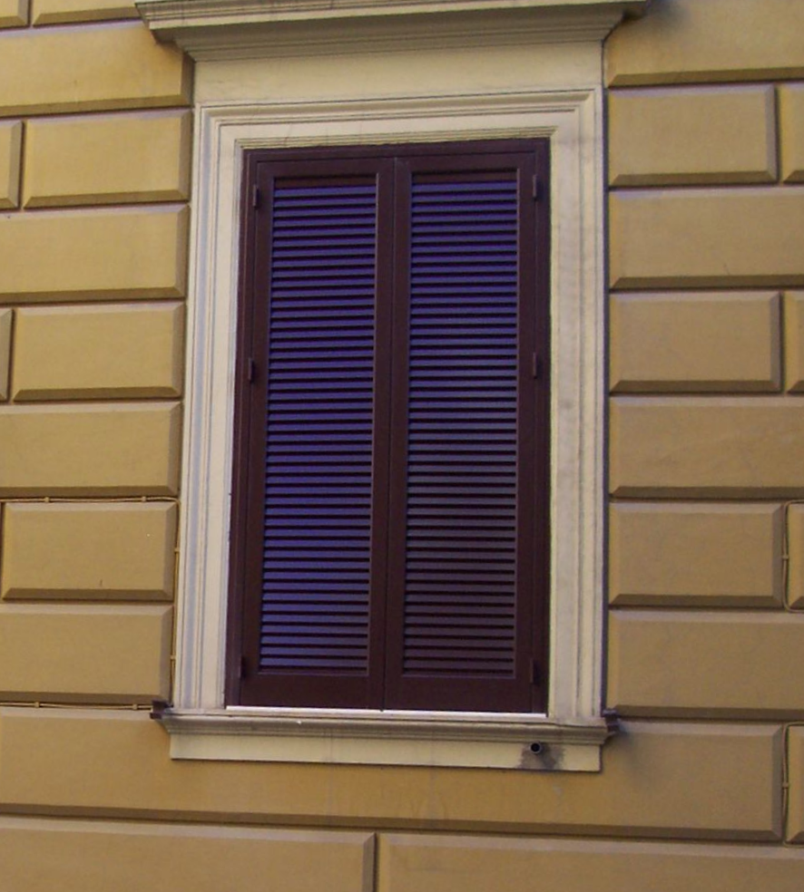 Una finestra con persiane chiuse a lamelle marrone scuro, incastonata in un muro di pietra beige con blocchi rettangolari in muratura.