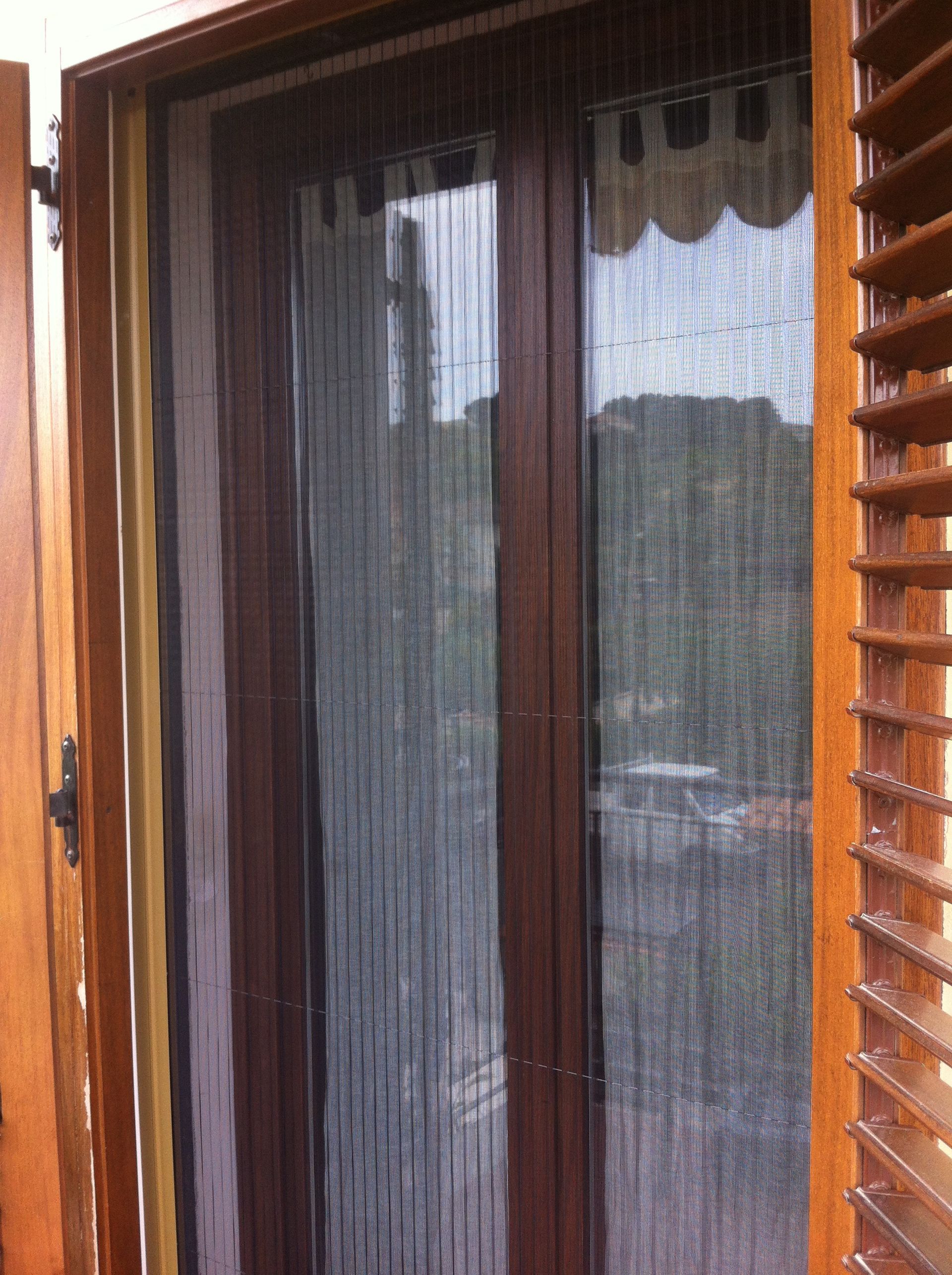 Un telaio di porta in legno marrone scuro con una zanzariera verticale a fili che pende sui vetri.