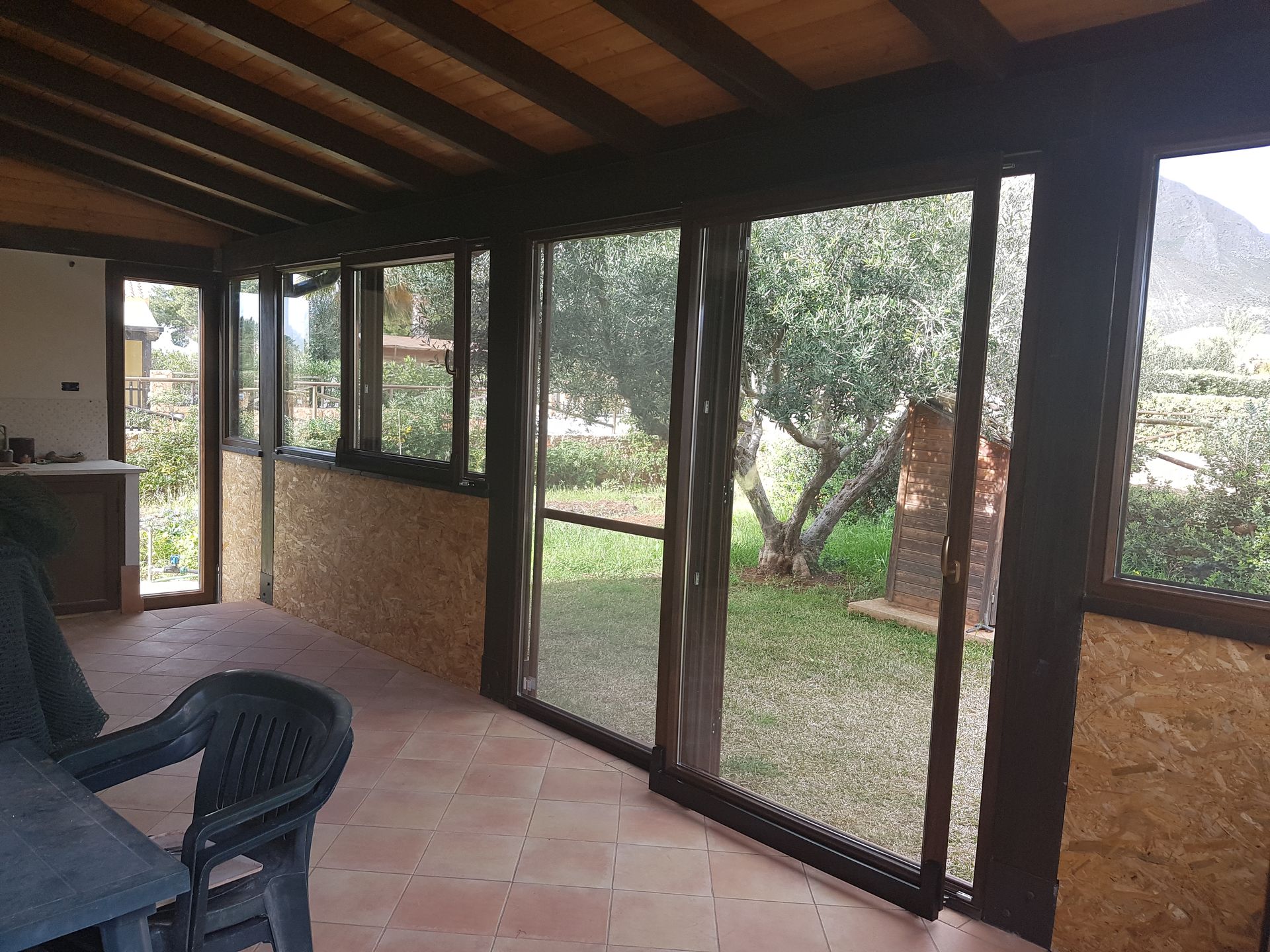 Un patio coperto con soffitto a travi in ​​legno, pavimento piastrellato, sedia in plastica e grandi finestre in vetro che si affacciano su un giardino.