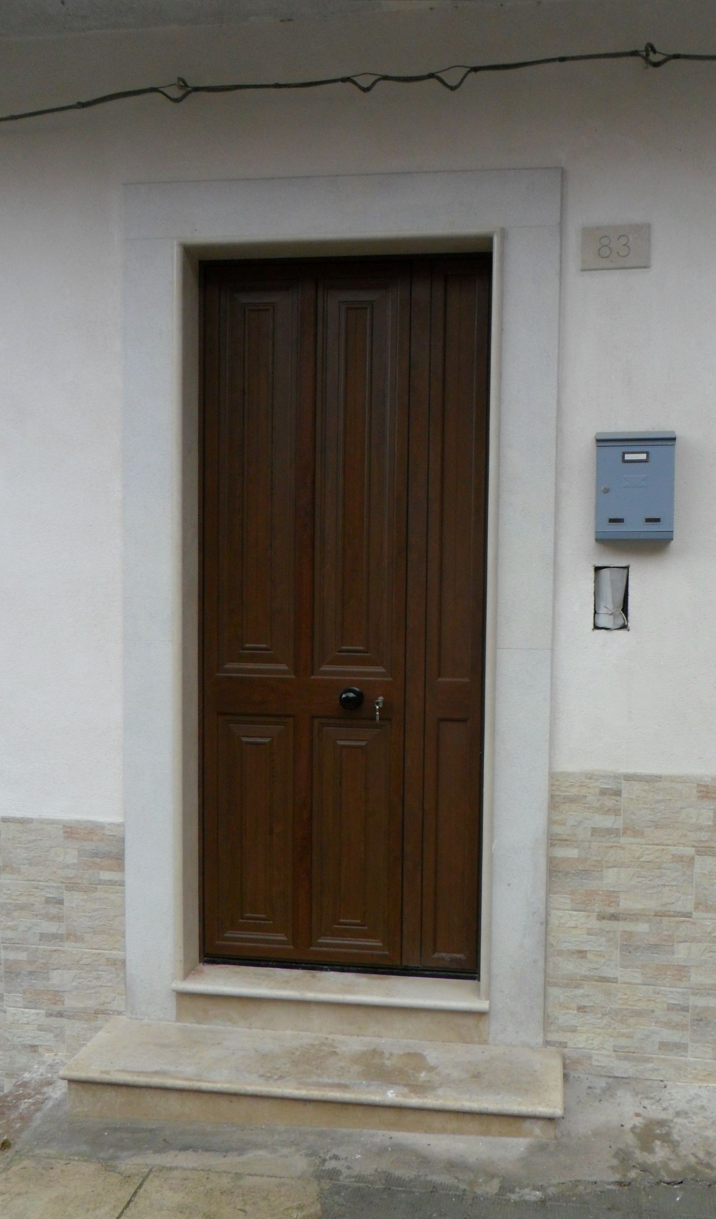 Una porta di legno marrone con pannelli in rilievo, incastonata in una cornice di pietra bianca con un gradino in pietra, su un muro bianco con una cassetta delle lettere.