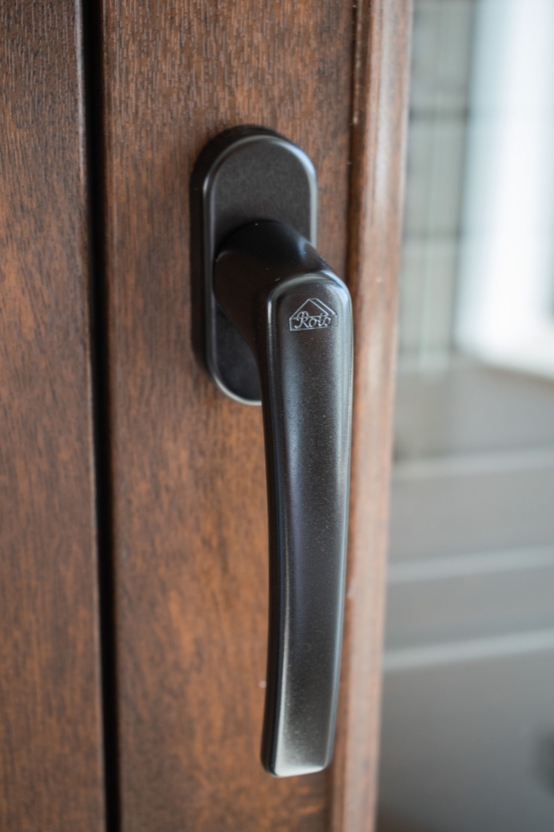 Una maniglia per finestra nera con un piccolo logo, montata su un telaio della finestra con finitura effetto legno marrone.