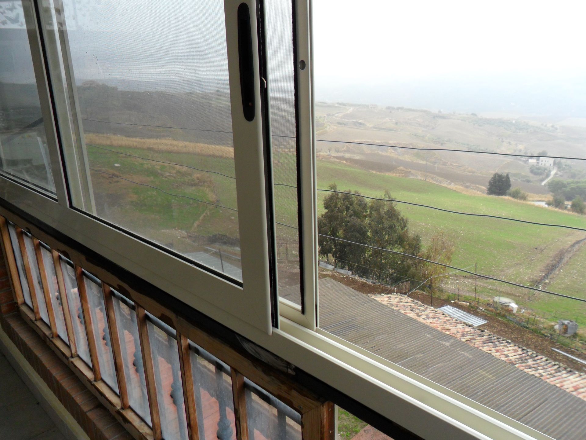 Una finestra scorrevole in vetro si affaccia su un paesaggio verdeggiante e collinare, con un tetto di tegole parzialmente visibile al di sotto.