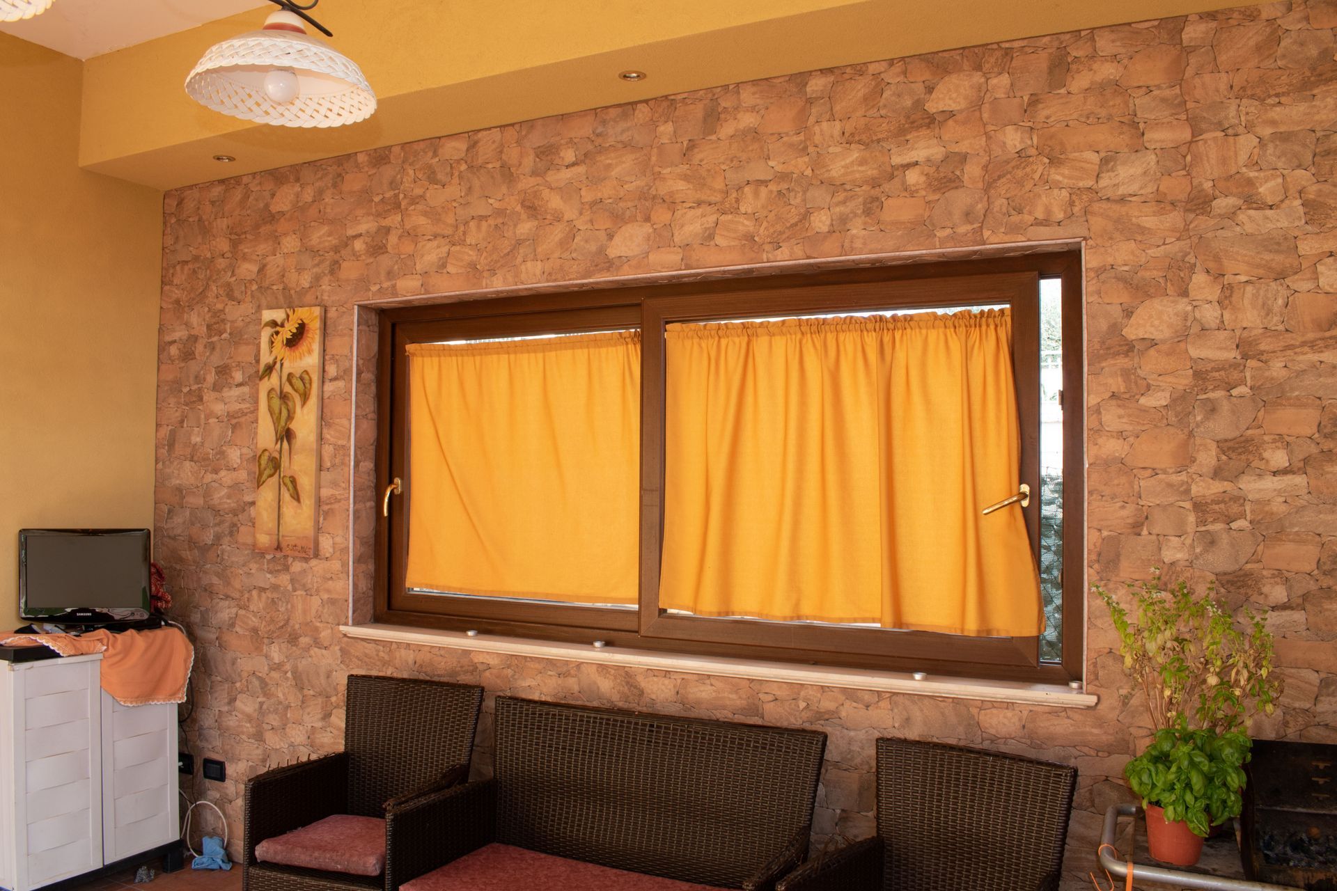 Uno spazio interno luminoso con pareti che imitano la pietra, tende gialle a una finestra, un divano e una pianta in vaso.
