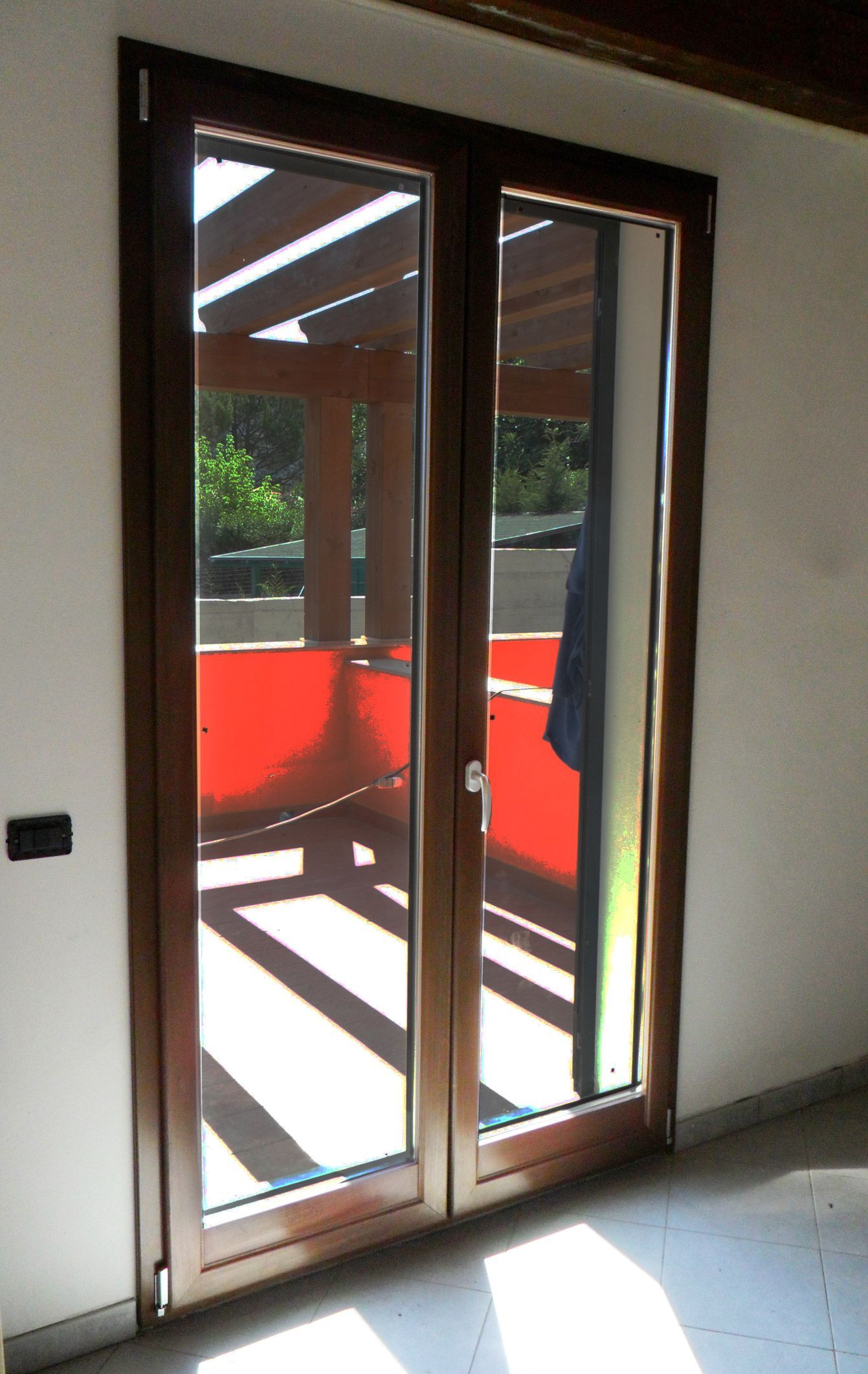 Una porta a doppio battente in vetro con cornice marrone conduce a un balcone con pareti color arancione acceso e ombre sul pavimento.