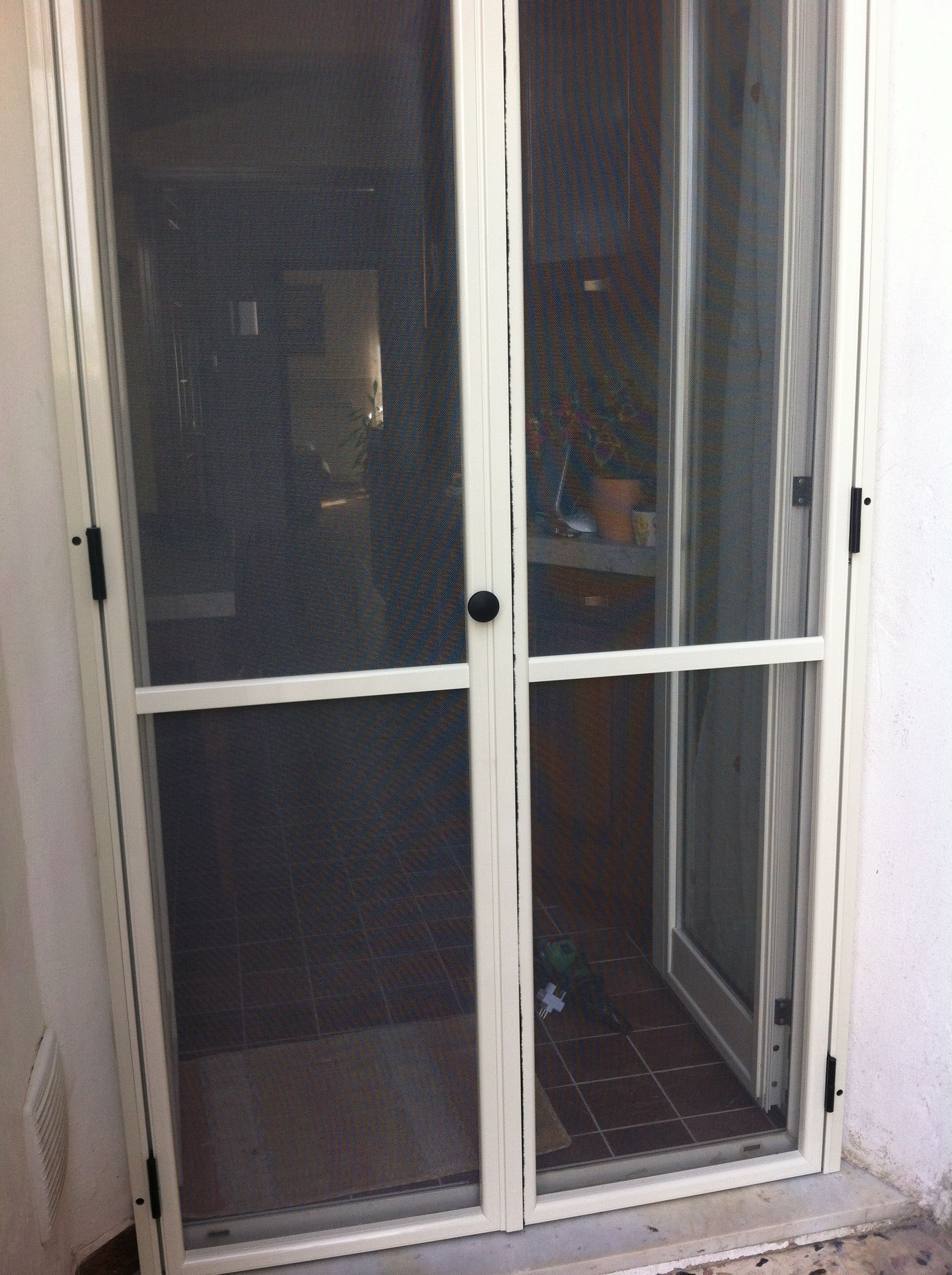 Doppie porte a zanzariera color crema installate in una porta, che lasciano intravedere una stanza interna scarsamente illuminata.