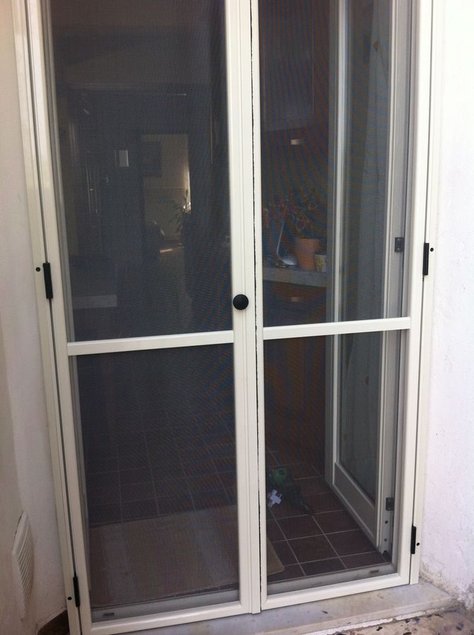 Doppie porte a zanzariera color crema installate in una porta, che lasciano intravedere una stanza interna scarsamente illuminata.