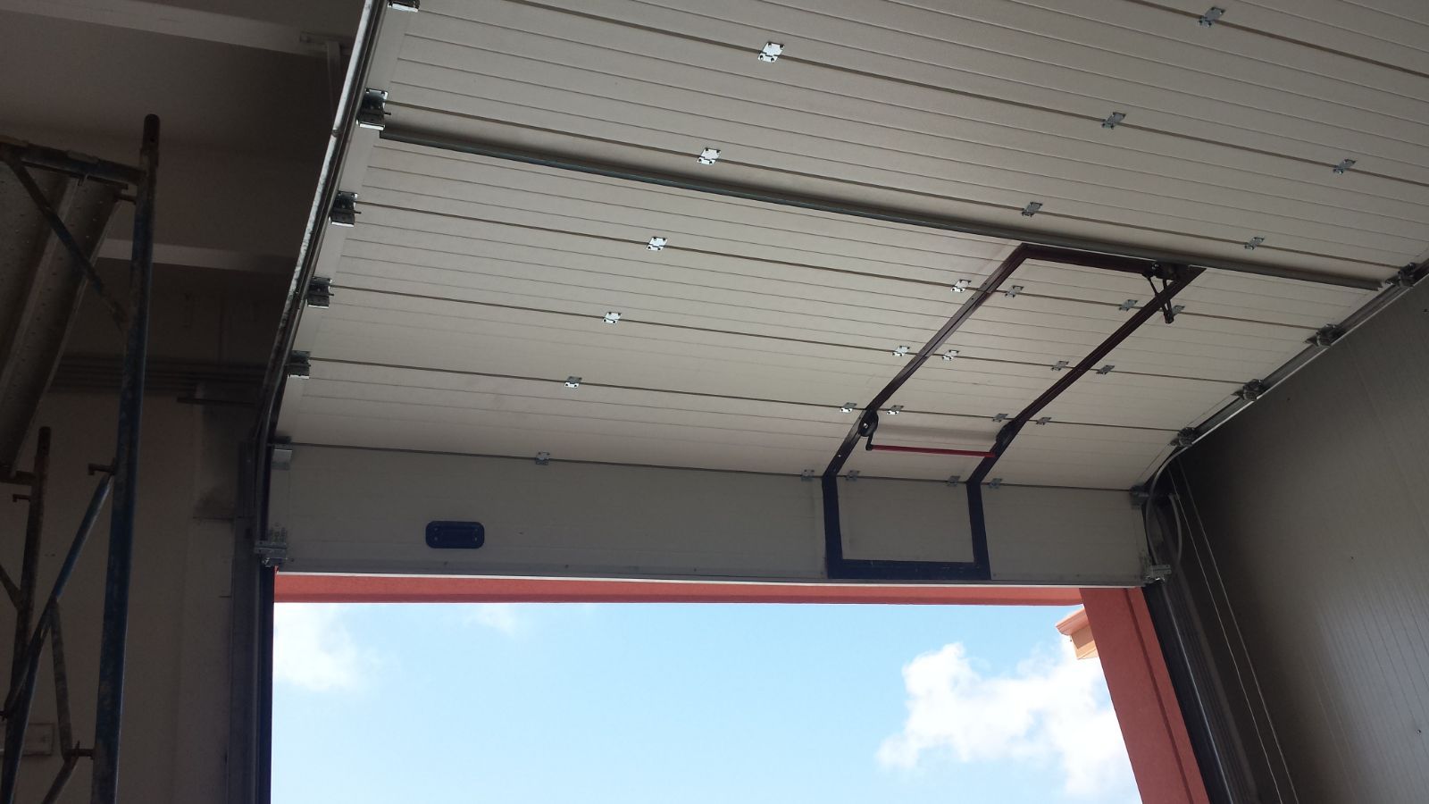 Una porta da garage sezionale bianca, parzialmente sollevata, lascia intravedere il cielo azzurro all'esterno.