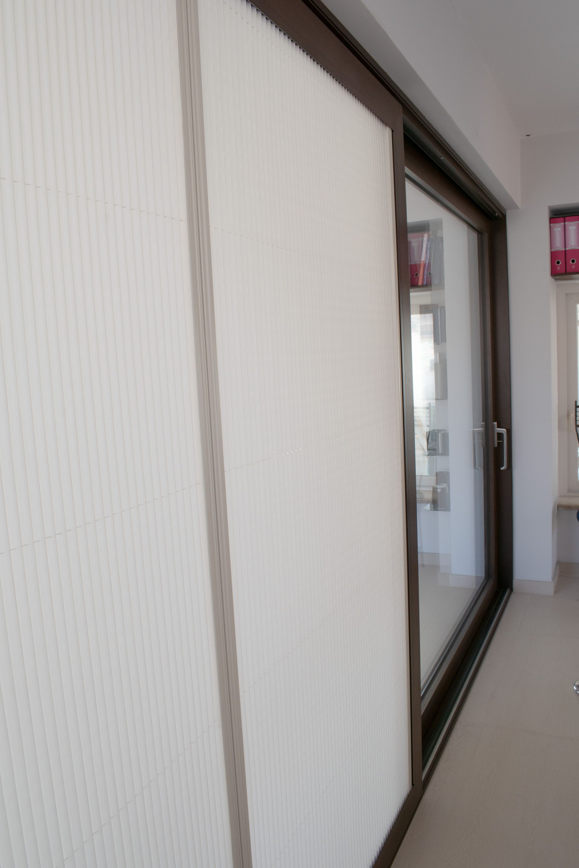 In una stanza ben illuminata, una porta a zanzariera bianca plissettata è parzialmente tirata su una porta scorrevole in vetro con cornice scura.