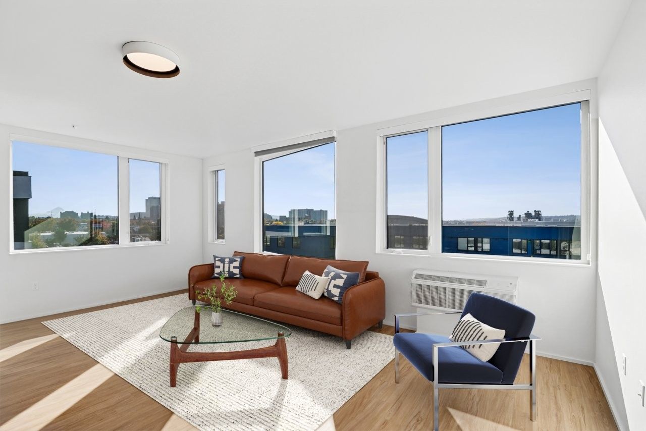 Bright living room with large windows, brown leather sofa, blue chair, and glass coffee table.