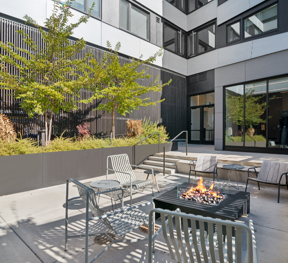Outdoor patio with chairs, a fire pit, and trees surrounded by a modern building.