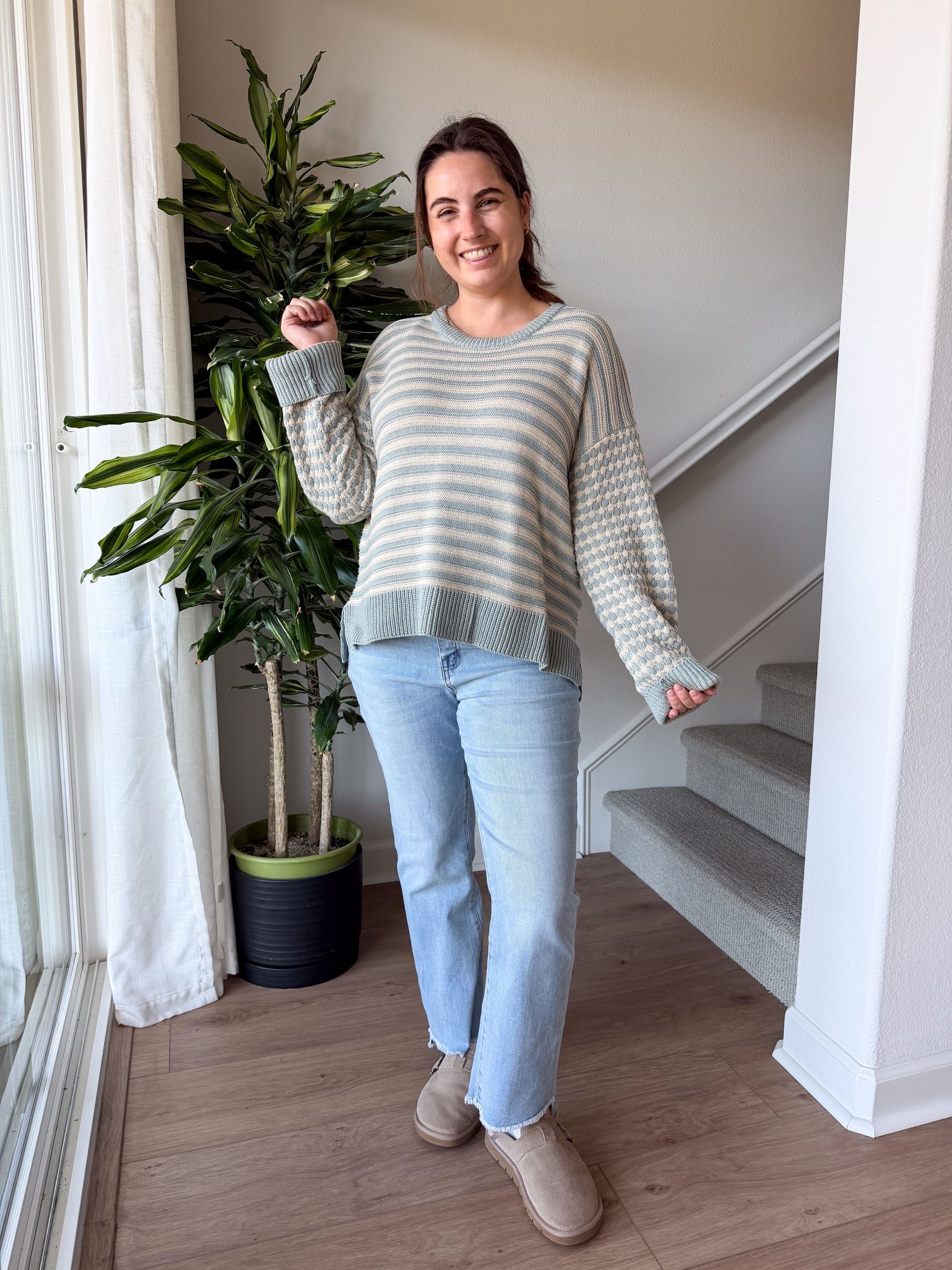 Woman in a sweater and jeans smiles near a staircase and potted plant.