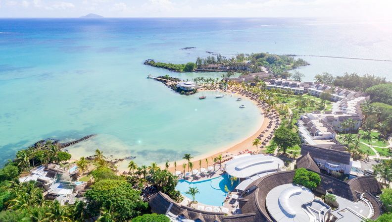 Aerial view of a tropical beach resort with turquoise water, a curved shoreline, pool, and palm trees.