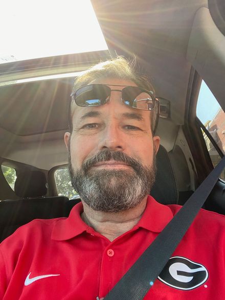 Man with beard and sunglasses in a car, wearing a red shirt with a logo, sunlight shining.