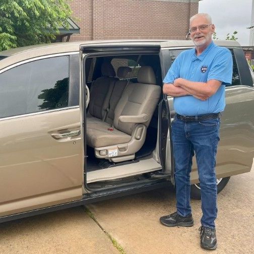 A man in a blue shirt is standing next to a minivan with the door open.