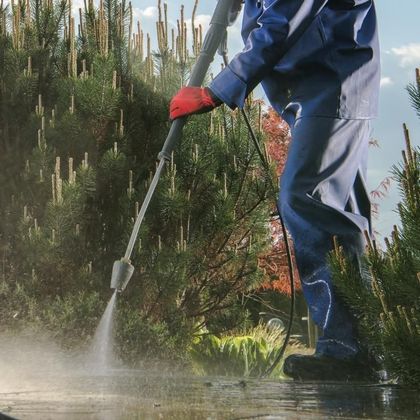 Person in blue suit pressure-washing concrete, wearing red gloves, with greenery in the background.