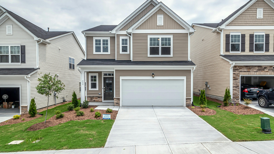 Two-story beige house with white garage door, flanked by similar townhomes, on a green lawn with concrete driveway.