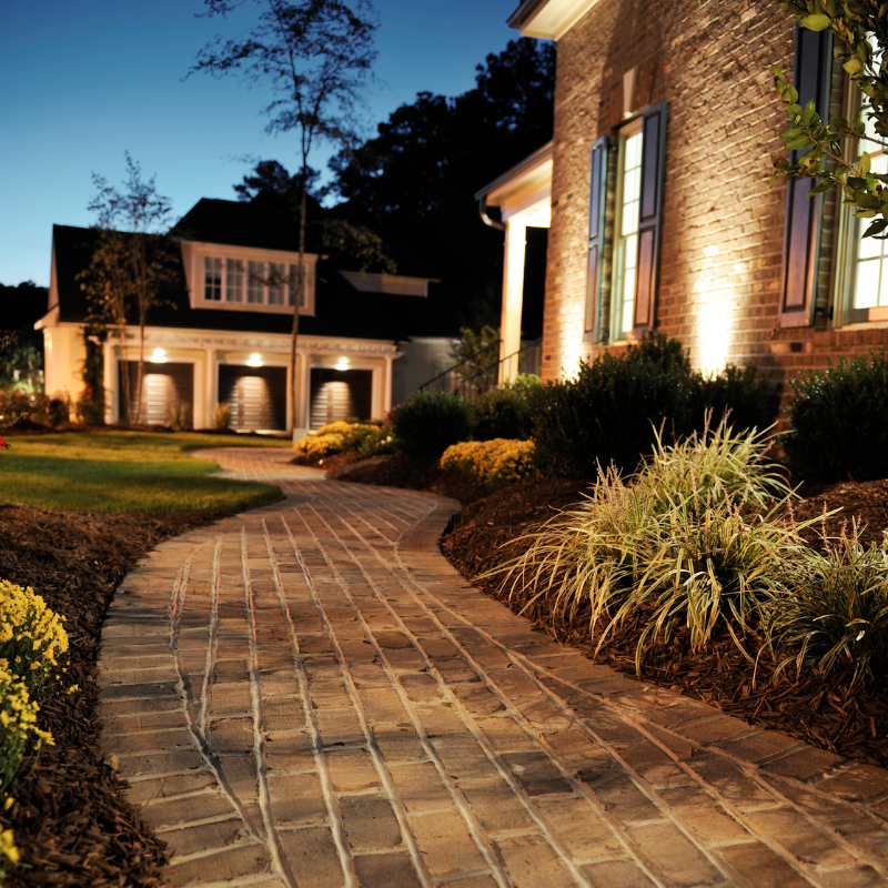 Brick path leads to a house with illuminated facade and a detached garage at dusk.