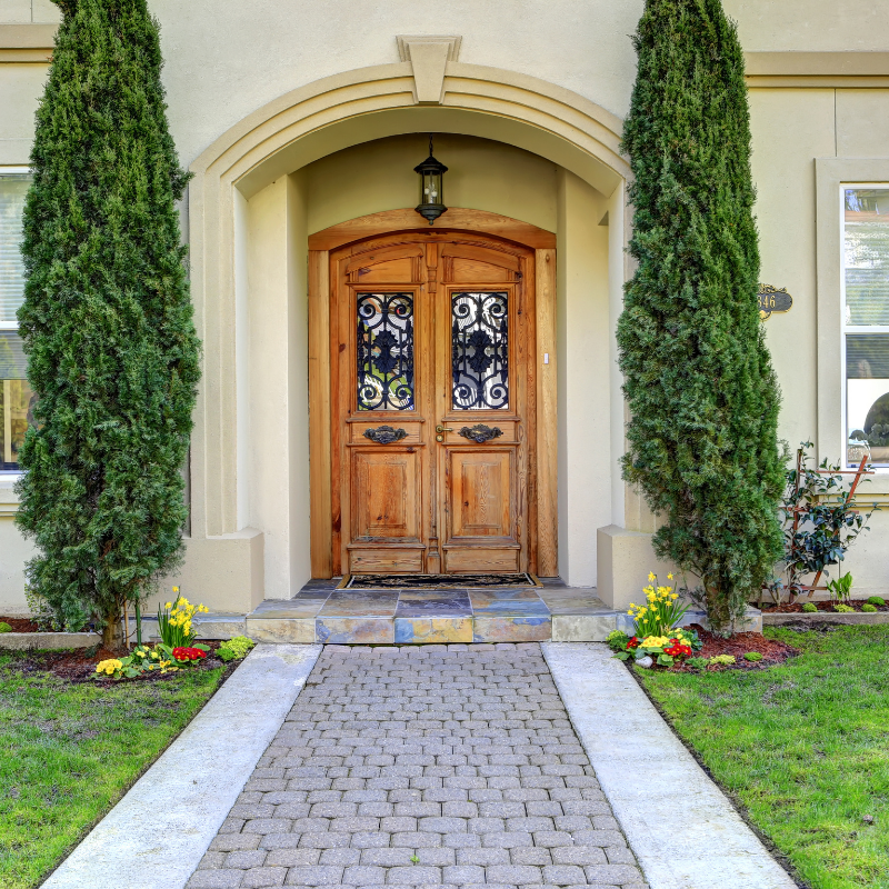 Wooden double doors with arched entryway, flanked by tall green trees, leading to a home with a brick path.