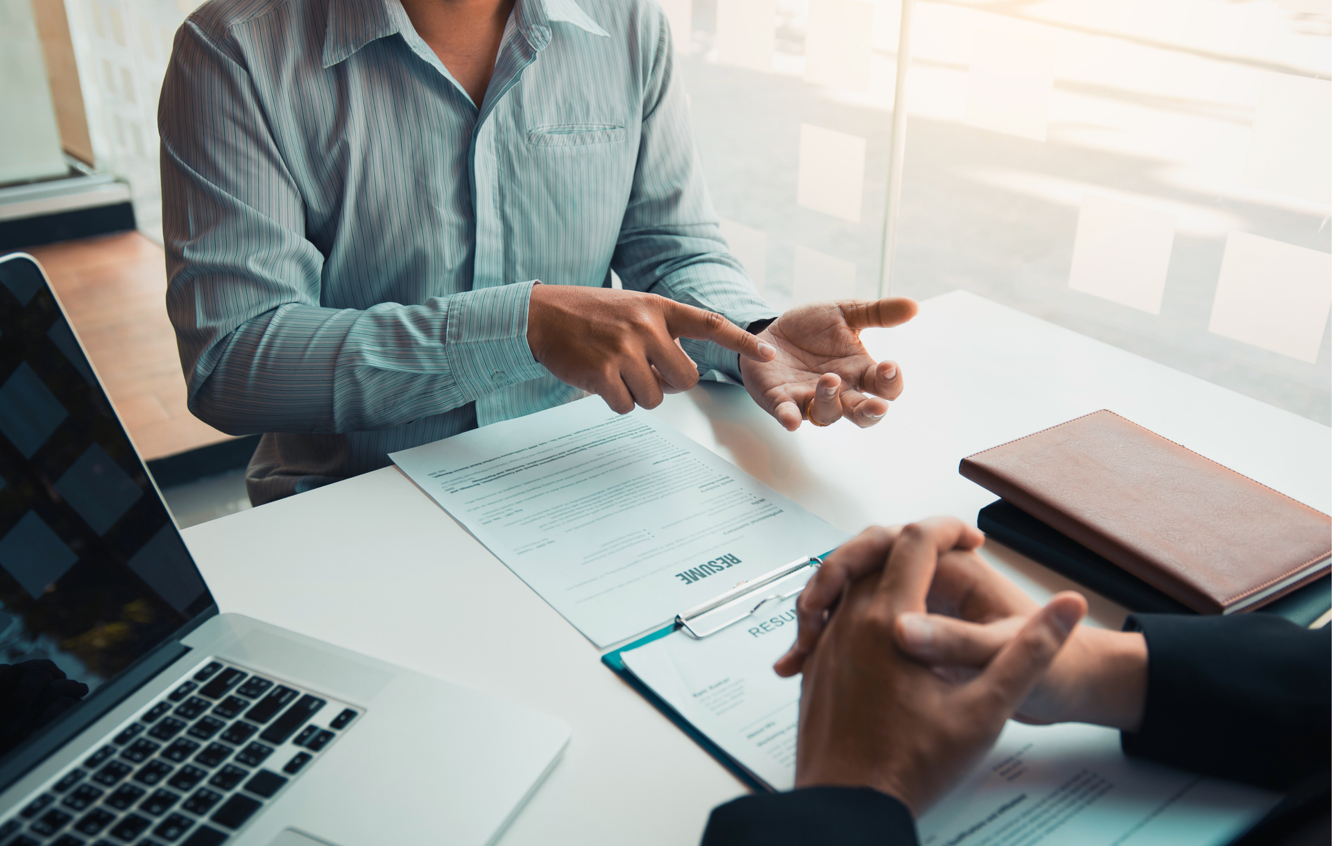 Two people in a professional setting discuss over a table with a laptop, resume, and notebook.