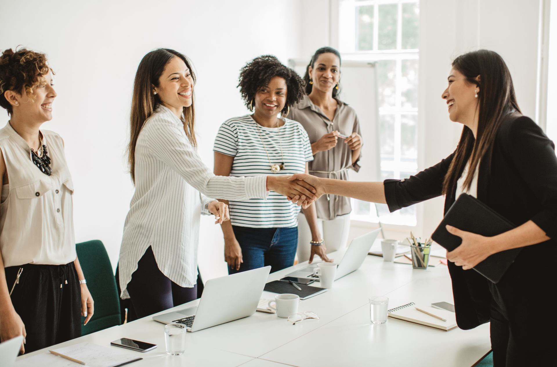 A group of women standing around a conference table as two women shake hands, smiling, in a meeting