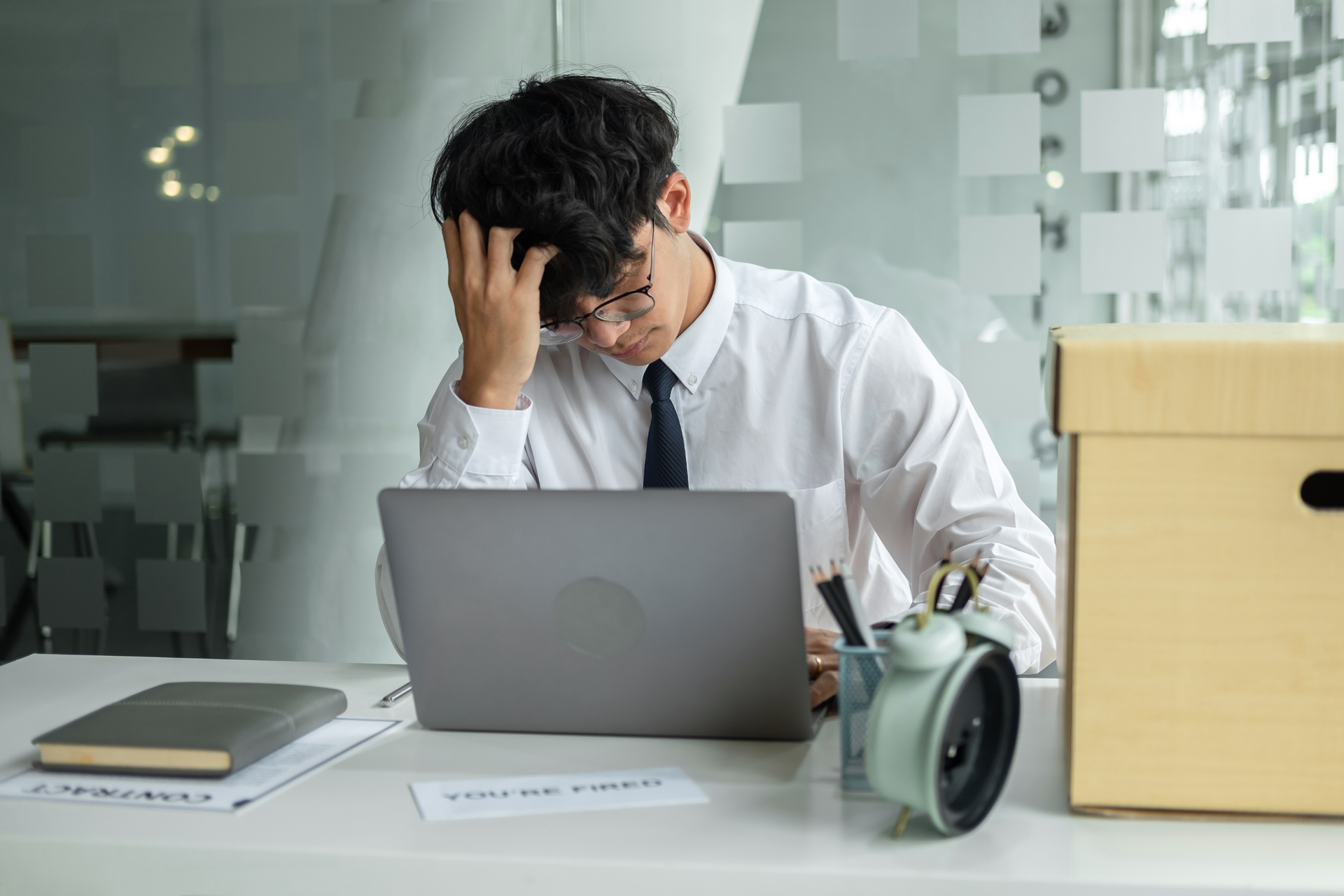 A man is sitting at a desk with a laptop and a termination letter. A cardboard box is beside him.