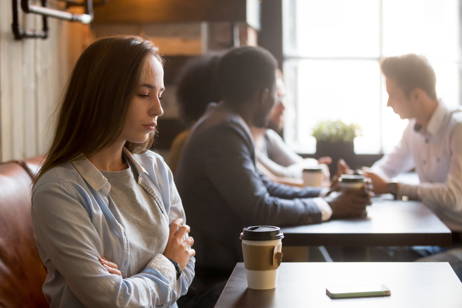 A woman sits alone in a coffee shop with a pensive expression, arms crossed. In the background, a group of people converse.