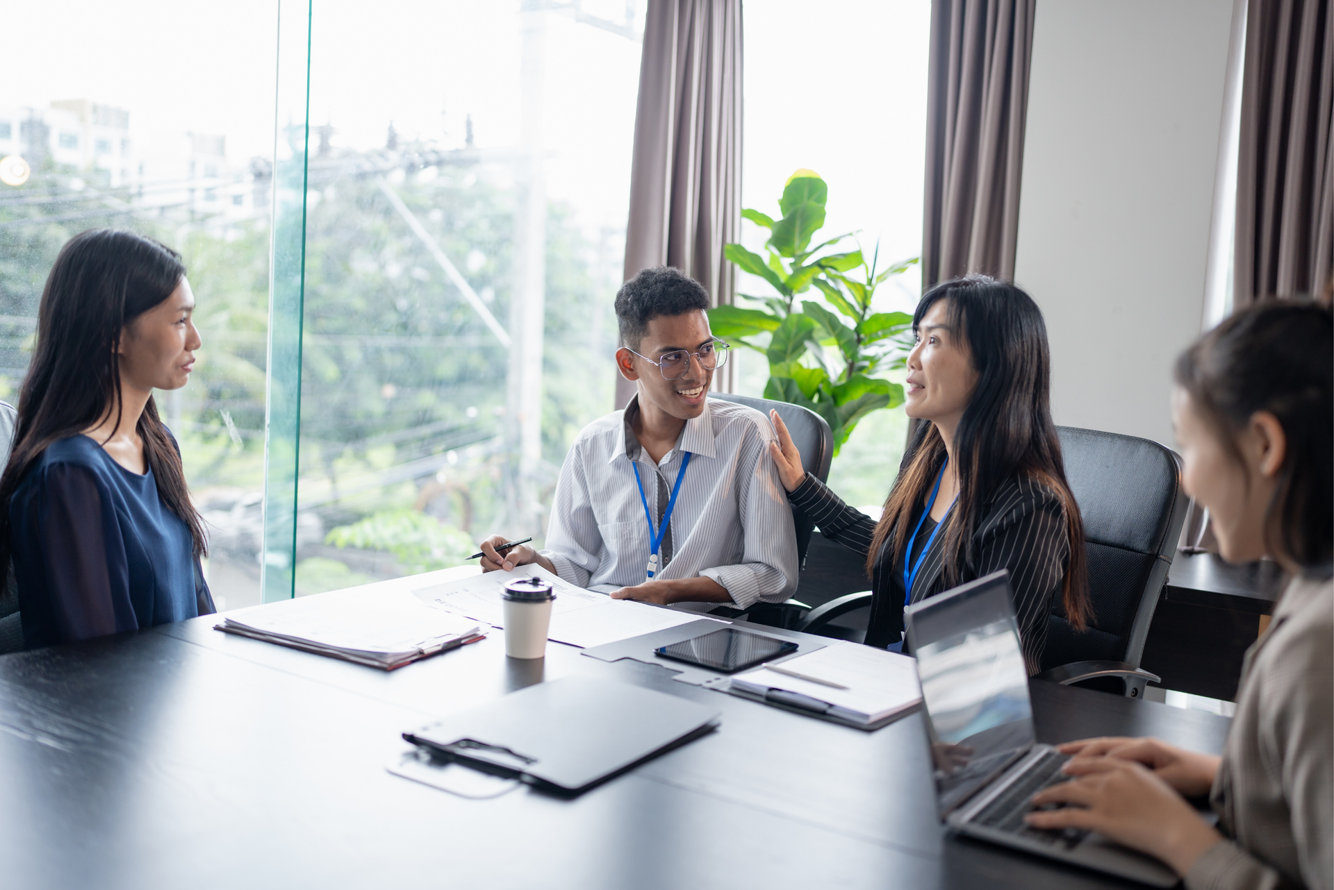 A group of four colleagues engage in a lively meeting around a table, with laptops and notes, in a bright office setting.