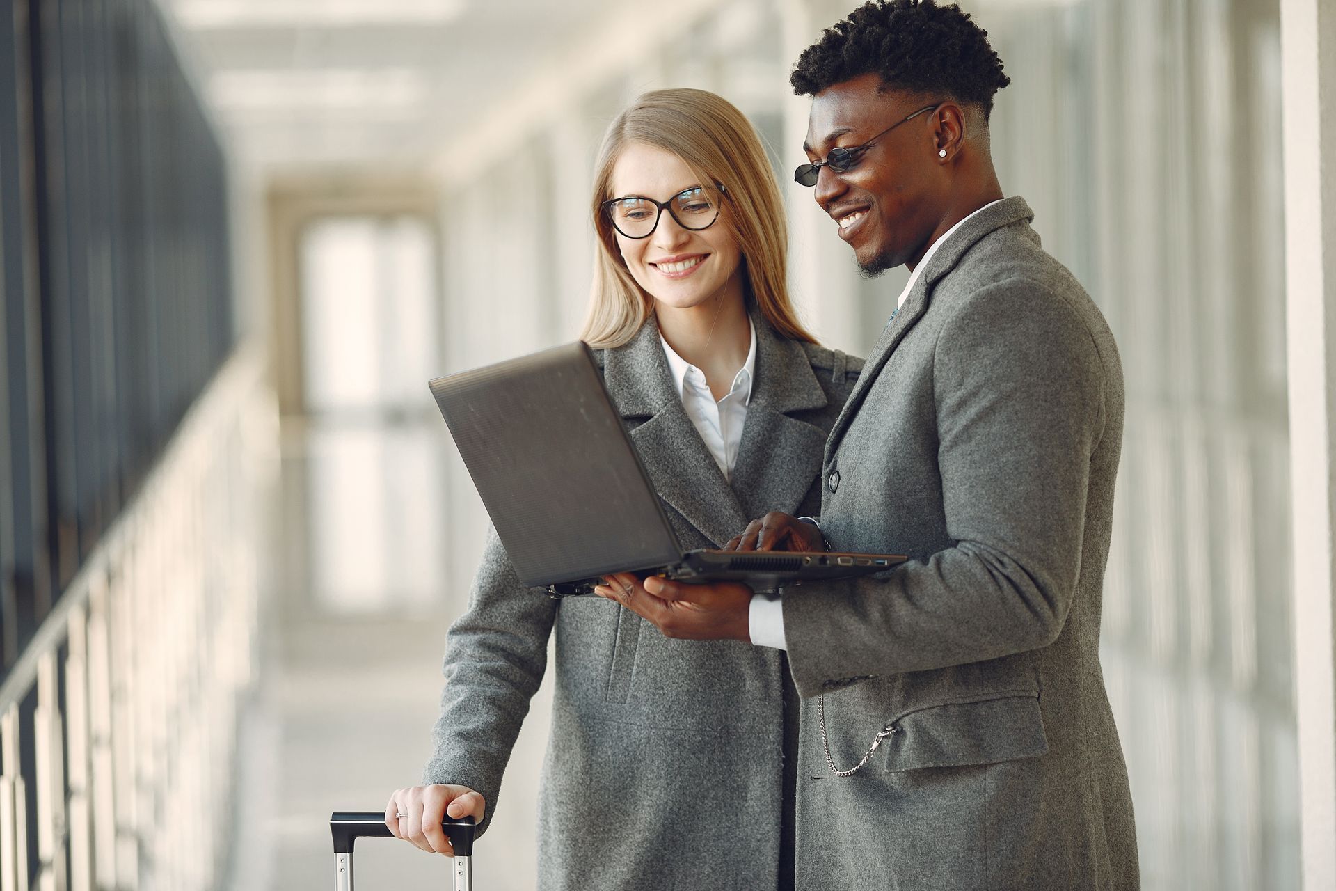 A man and a woman, both in gray coats, smile while looking at a laptop. The woman pulls a suitcase, suggesting travel.