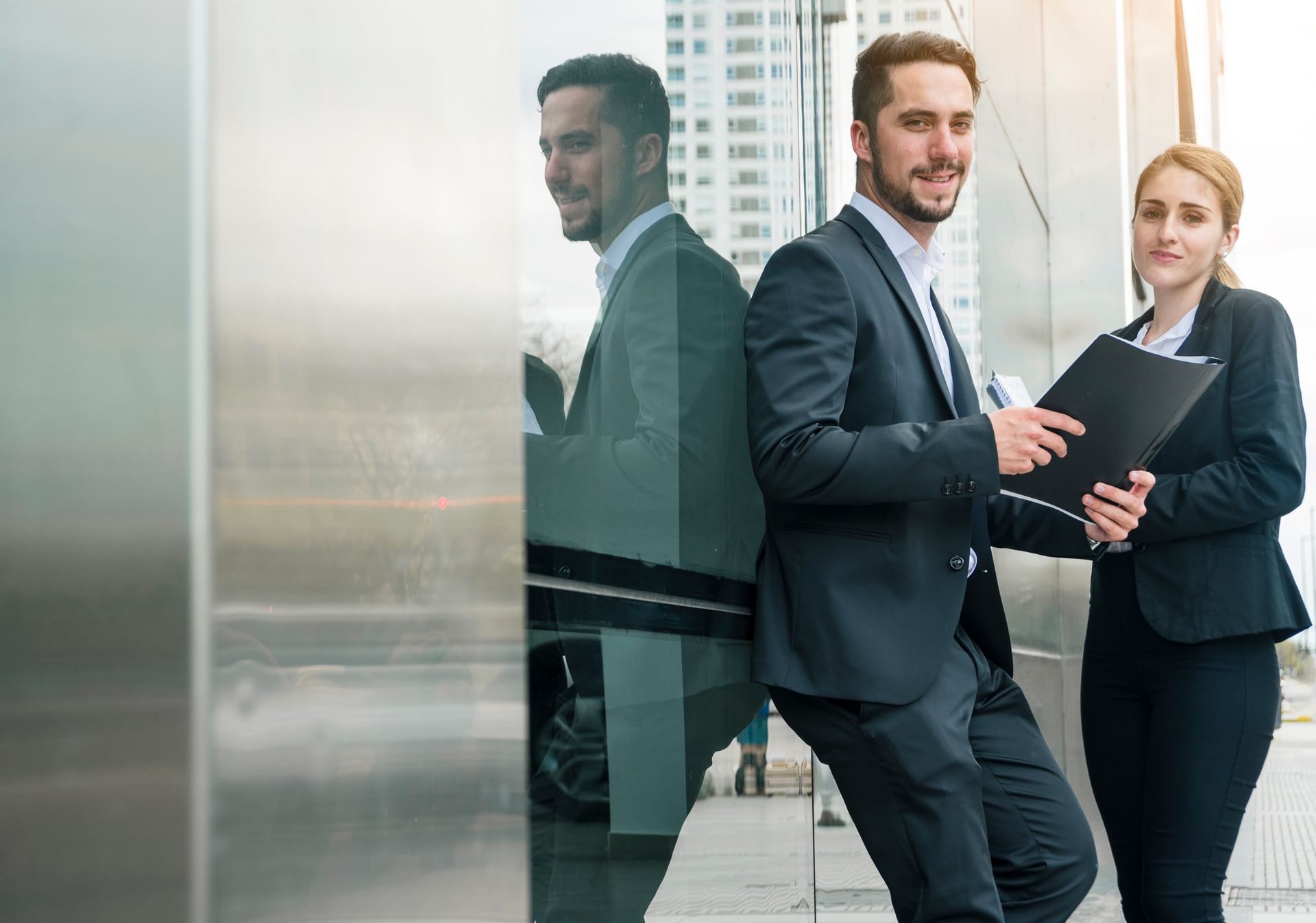 A man and woman in business attire stand outside a modern building. The man, holding a tablet, leans against a reflective glass wall, smiling. The woman holds a folder, exuding a professional demeanor, with city buildings in the background.