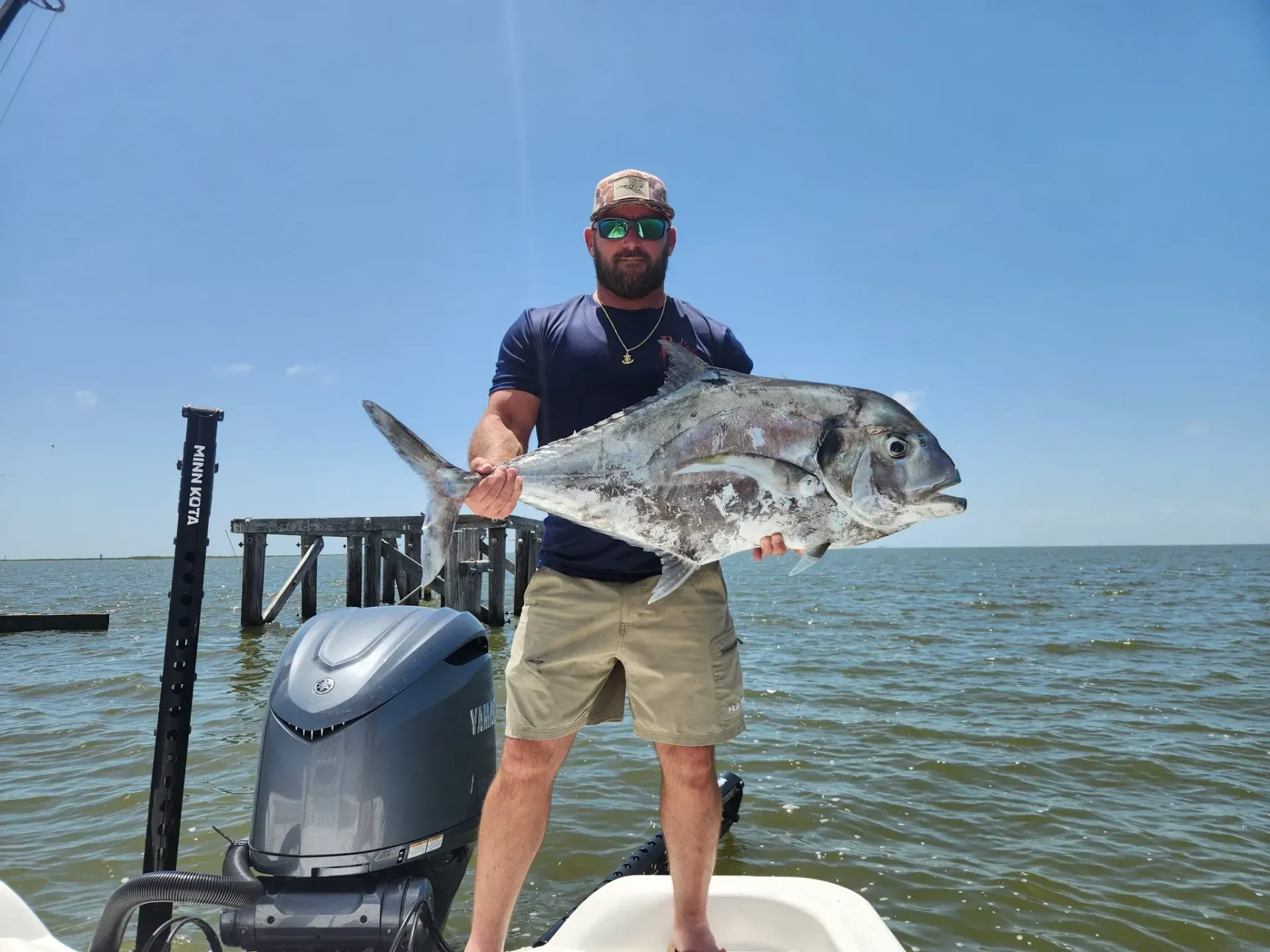 A person on a boat holding a large, silver fish, with a motor and a wooden pier in the background on a sunny day.