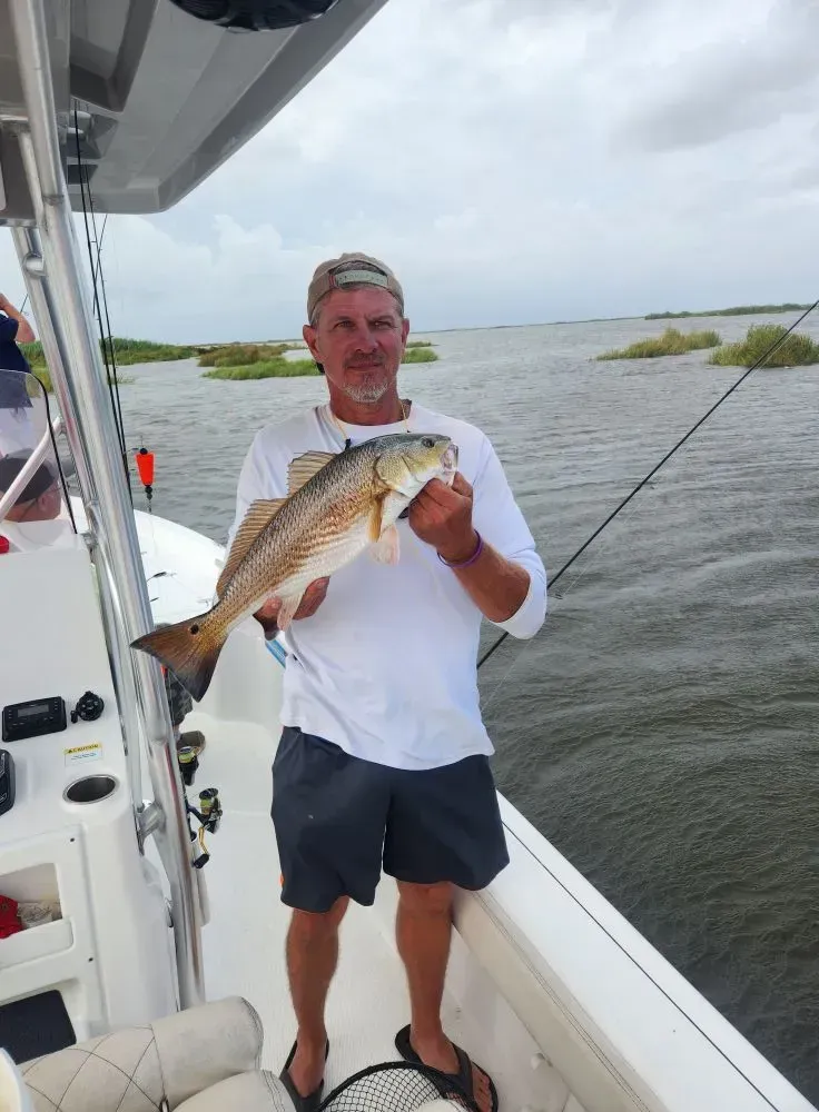 A person stands on a boat, holding a caught redfish in front of them with both hands against a backdrop of water and marsh.