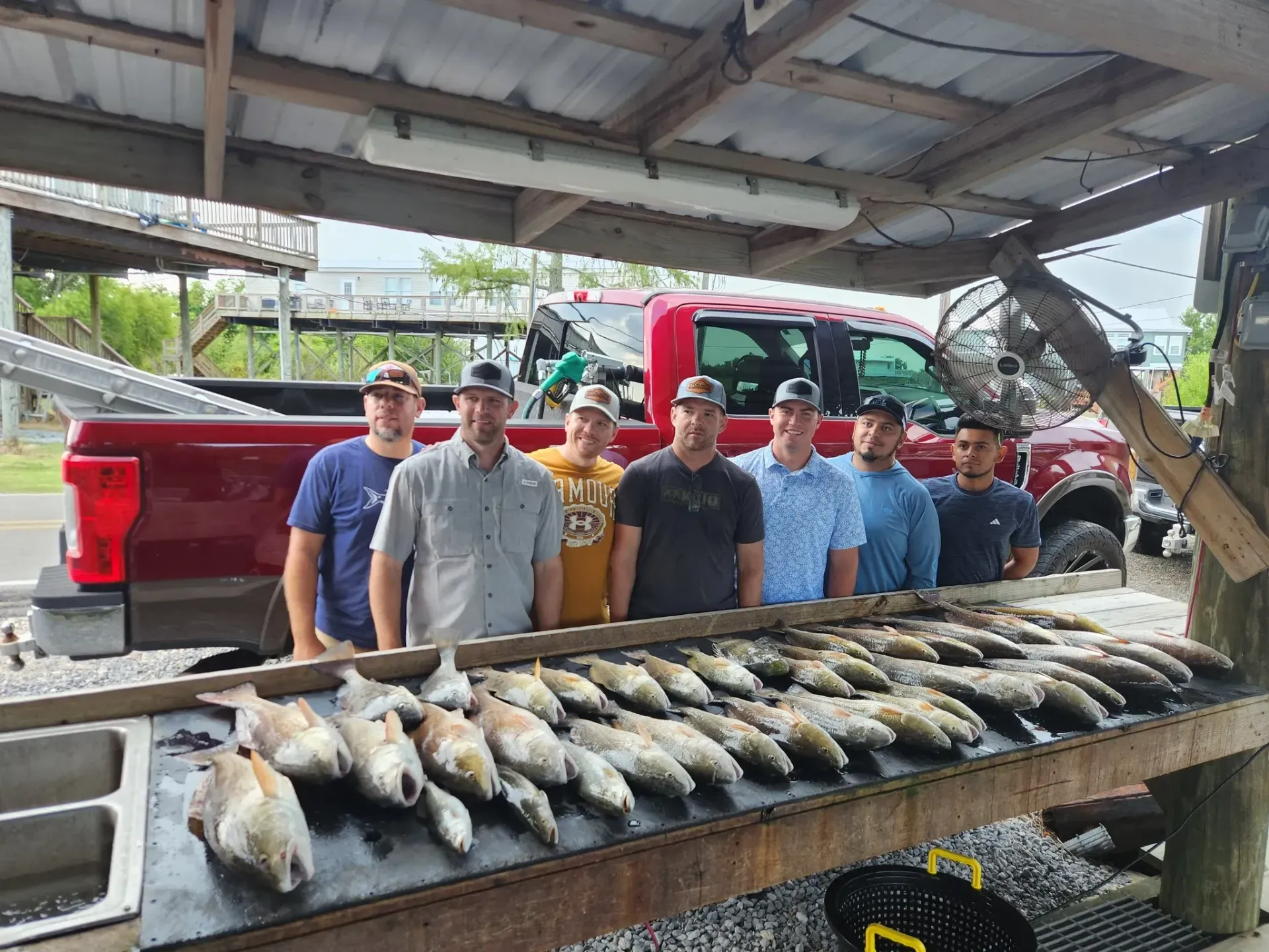 Seven people stand in a row behind a cleaning table covered with a large catch of fish, with a red pickup truck parked behind.